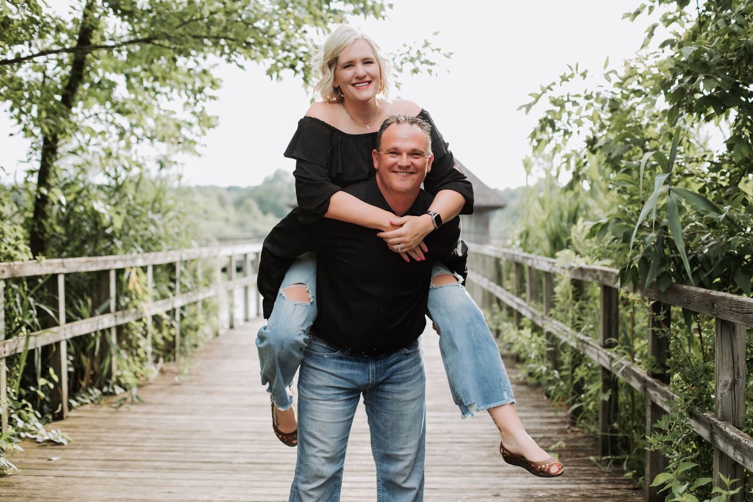 Man giving woman a piggyback ride on a wooden bridge surrounded by lush greenery.