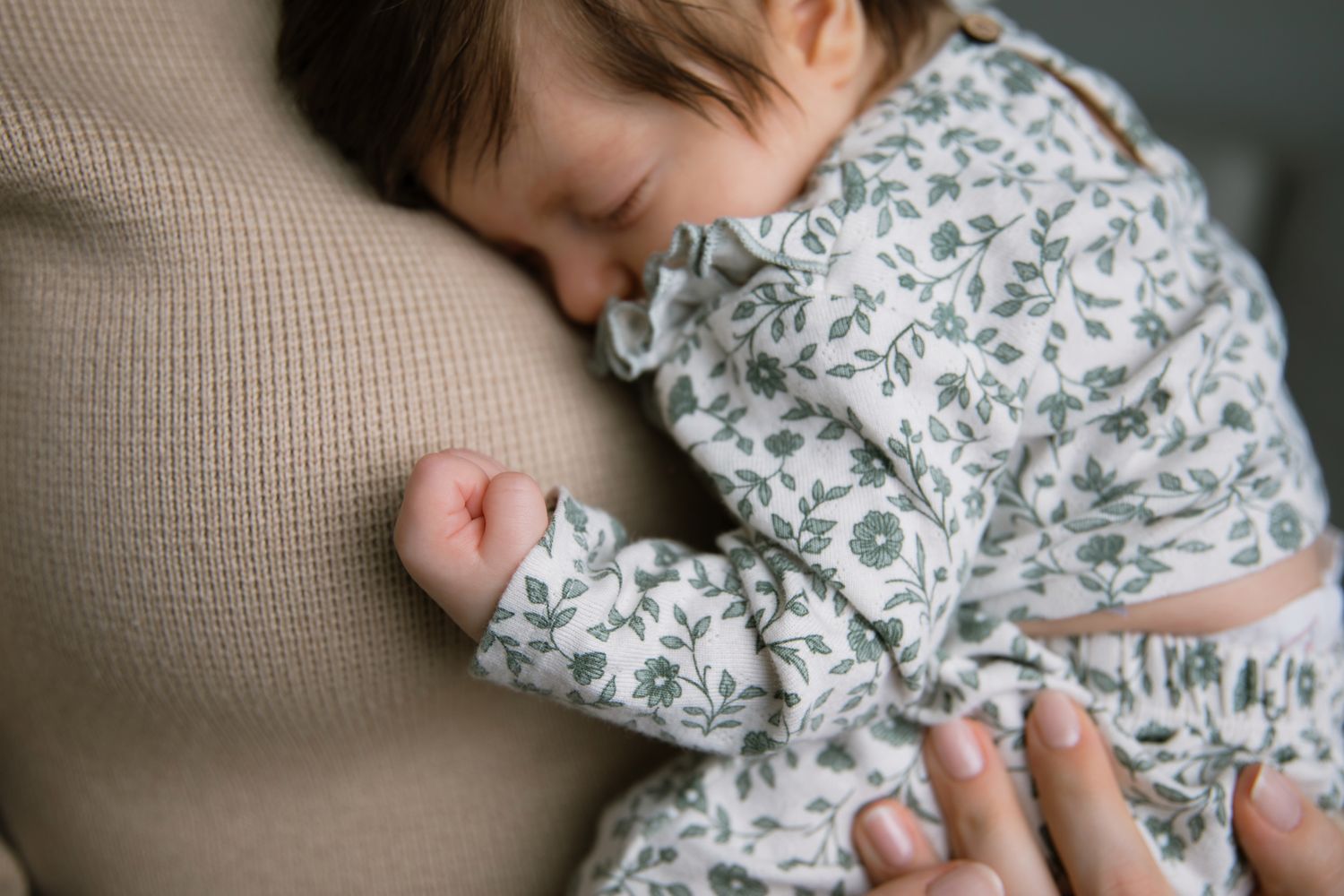 Sleeping newborn baby in floral outfit nestled against mothers shoulder.