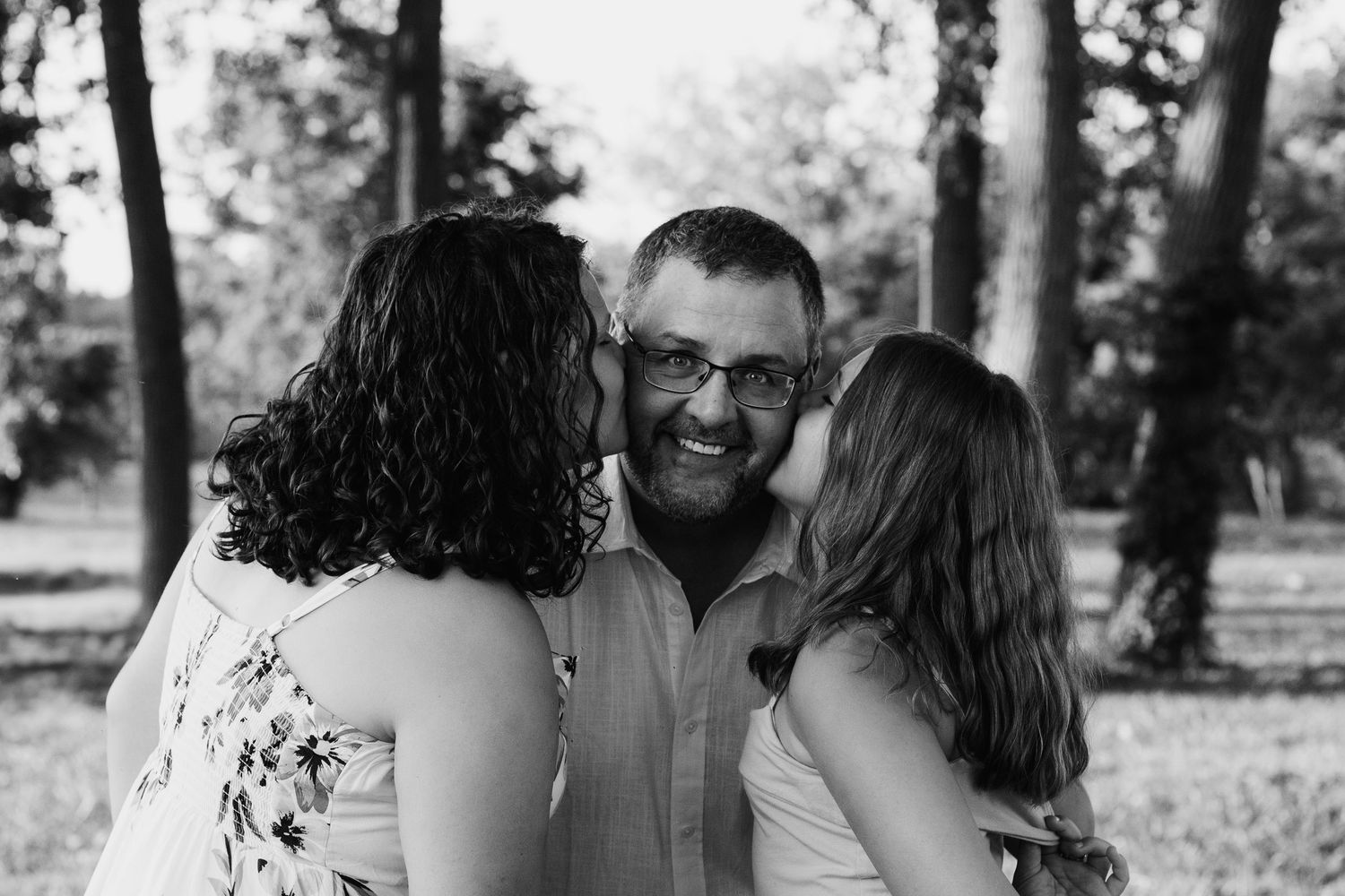 Man smiling as two women kiss his cheeks in black and white outdoor portrait.