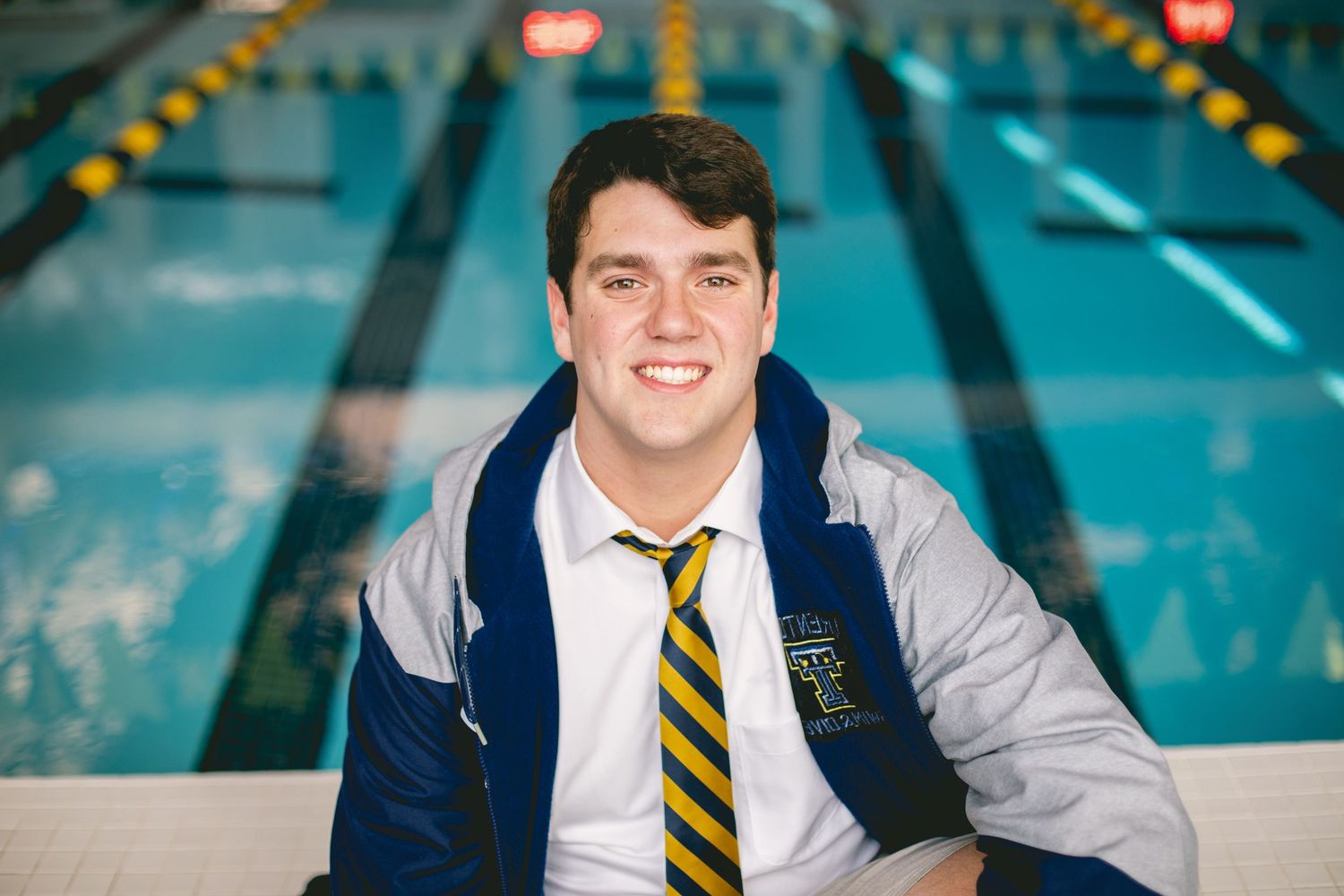 Young man in blue and gold varsity jacket and striped tie smiling poolside at an indoor swimming facility.