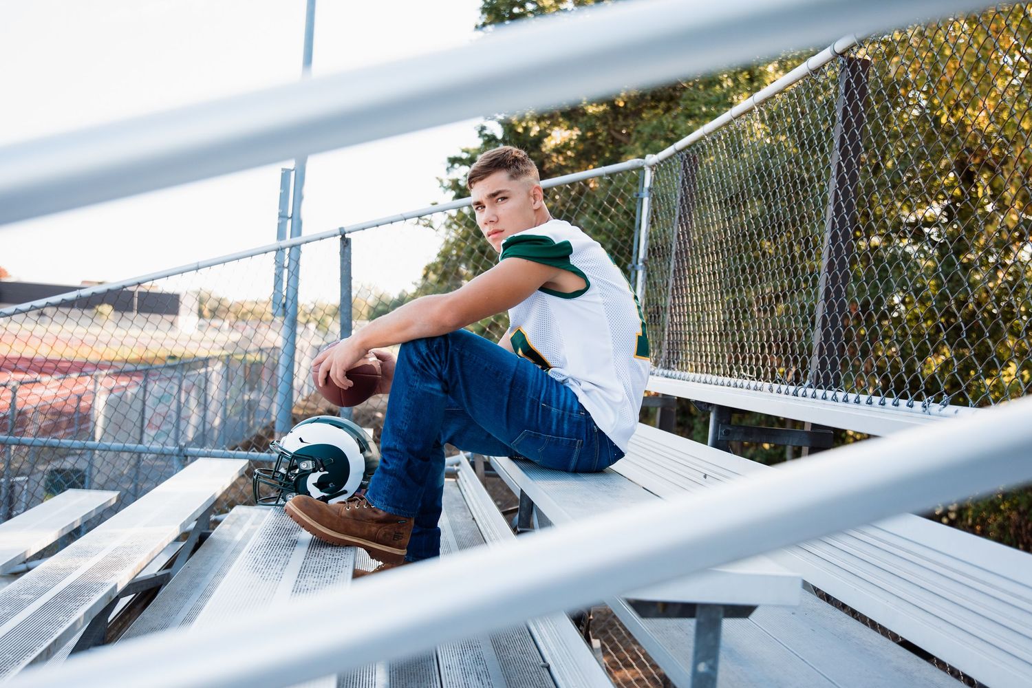 Young man in white shirt and jeans sits on bleachers holding a football helmet outdoors.