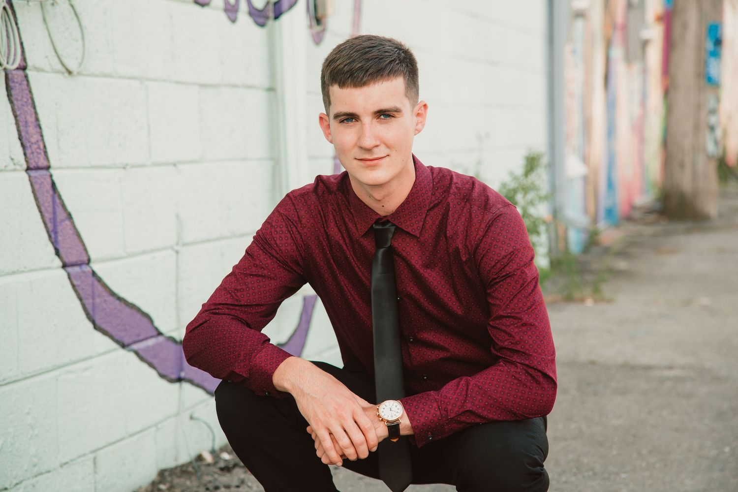 Young man in burgundy shirt and black tie crouching near graffiti wall, wearing a watch, in an urban alley setting.