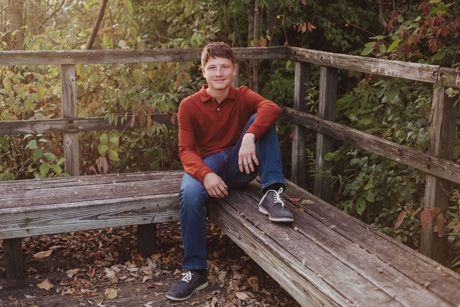 Teenage boy in red polo and jeans sitting relaxed on a wooden bench in an autumn garden.