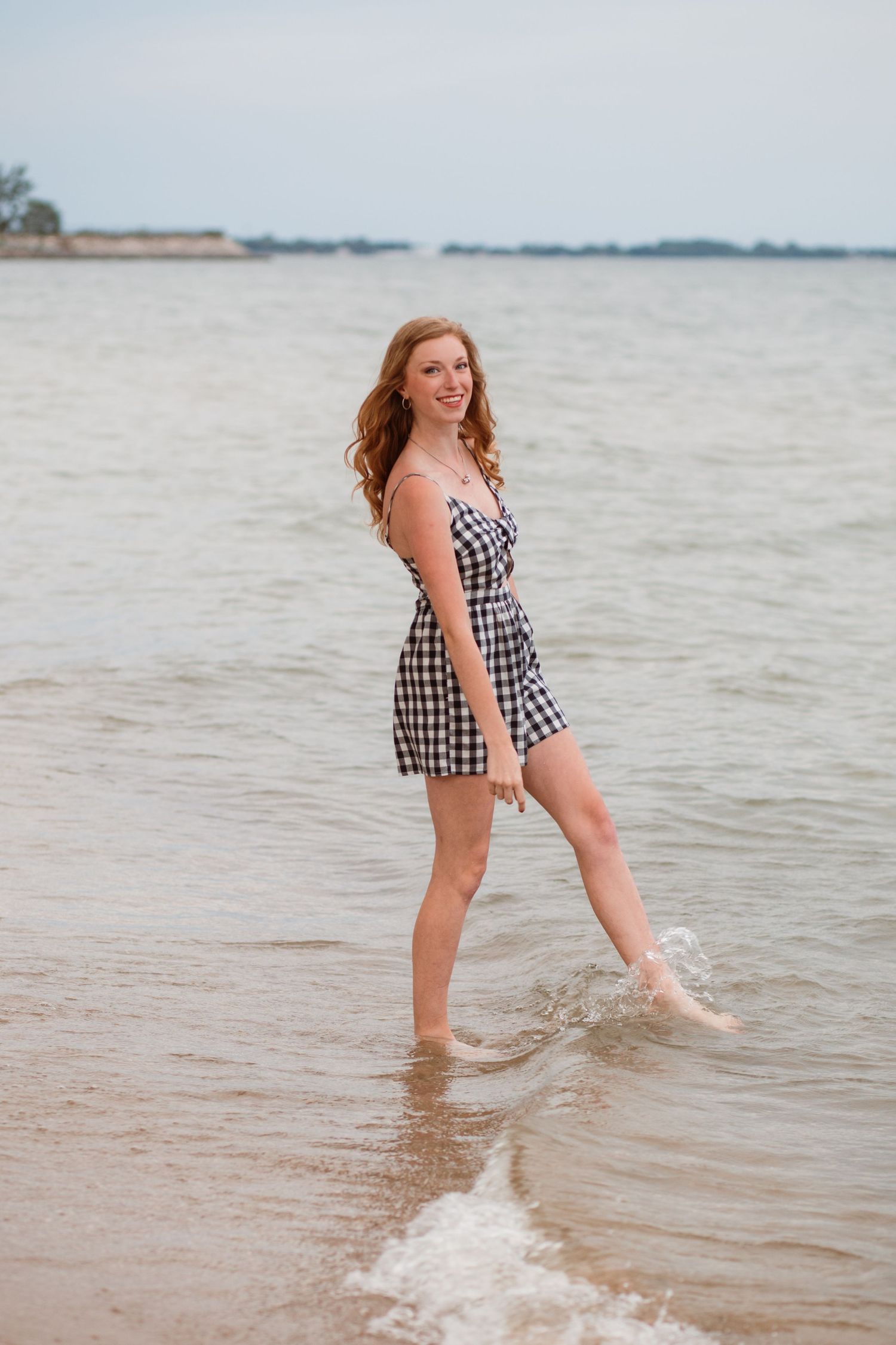 Young woman in gingham dress splashing in ocean waves on sandy beach smiling.