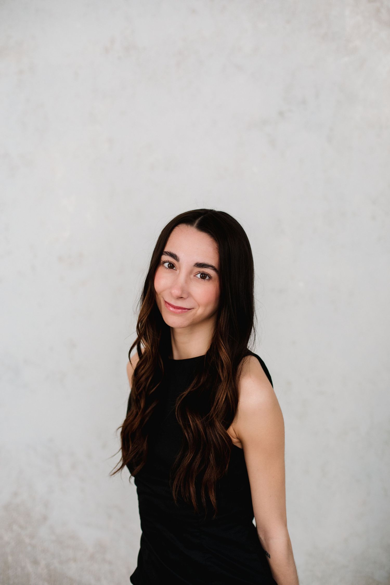A young woman with long dark hair in a black sleeveless dress smiles softly against a light gray textured wall.