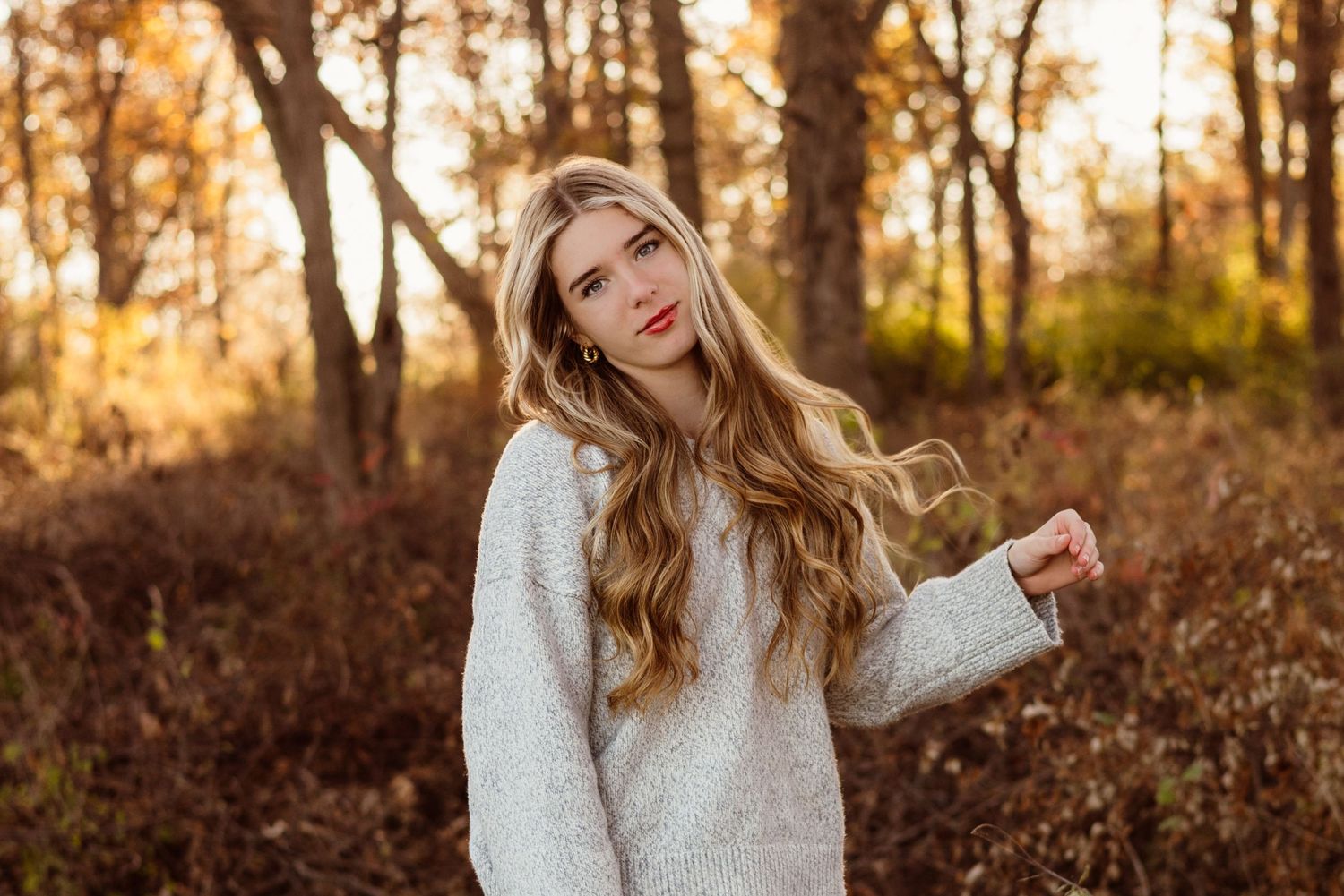 Blonde woman in white fuzzy sweater poses among autumn foliage in golden sunset light.