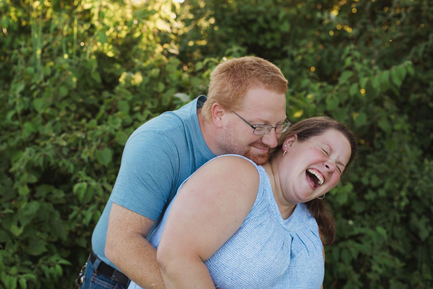 A joyful couple laughs together outdoors, with a red-haired man hugging a woman from behind near green foliage.