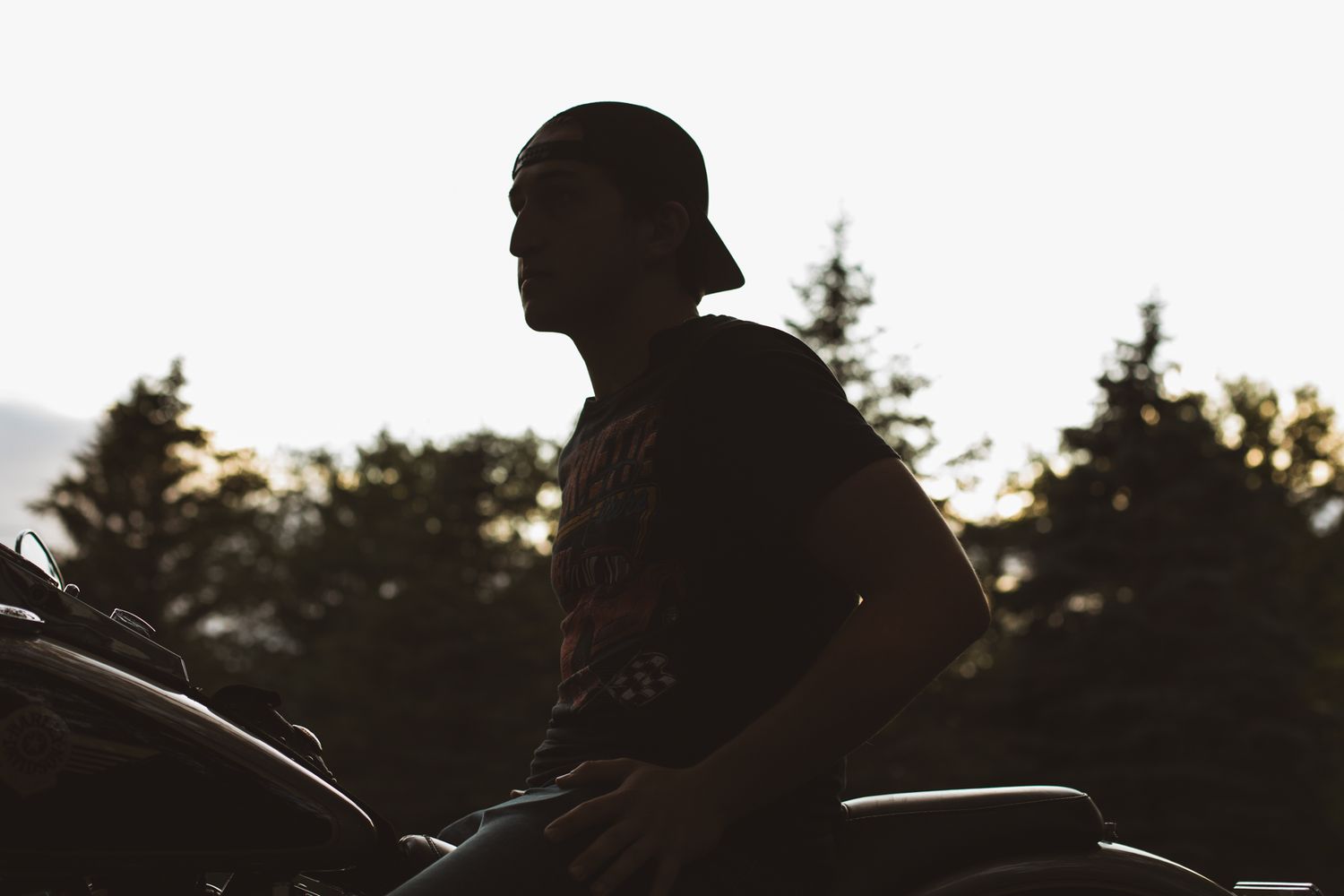 Silhouette of a man sitting on a motorcycle at sunset with trees in the background.