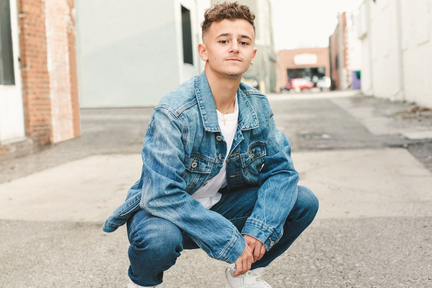 Young man in denim jacket and jeans crouching on an urban alley street in a casual fashion pose.