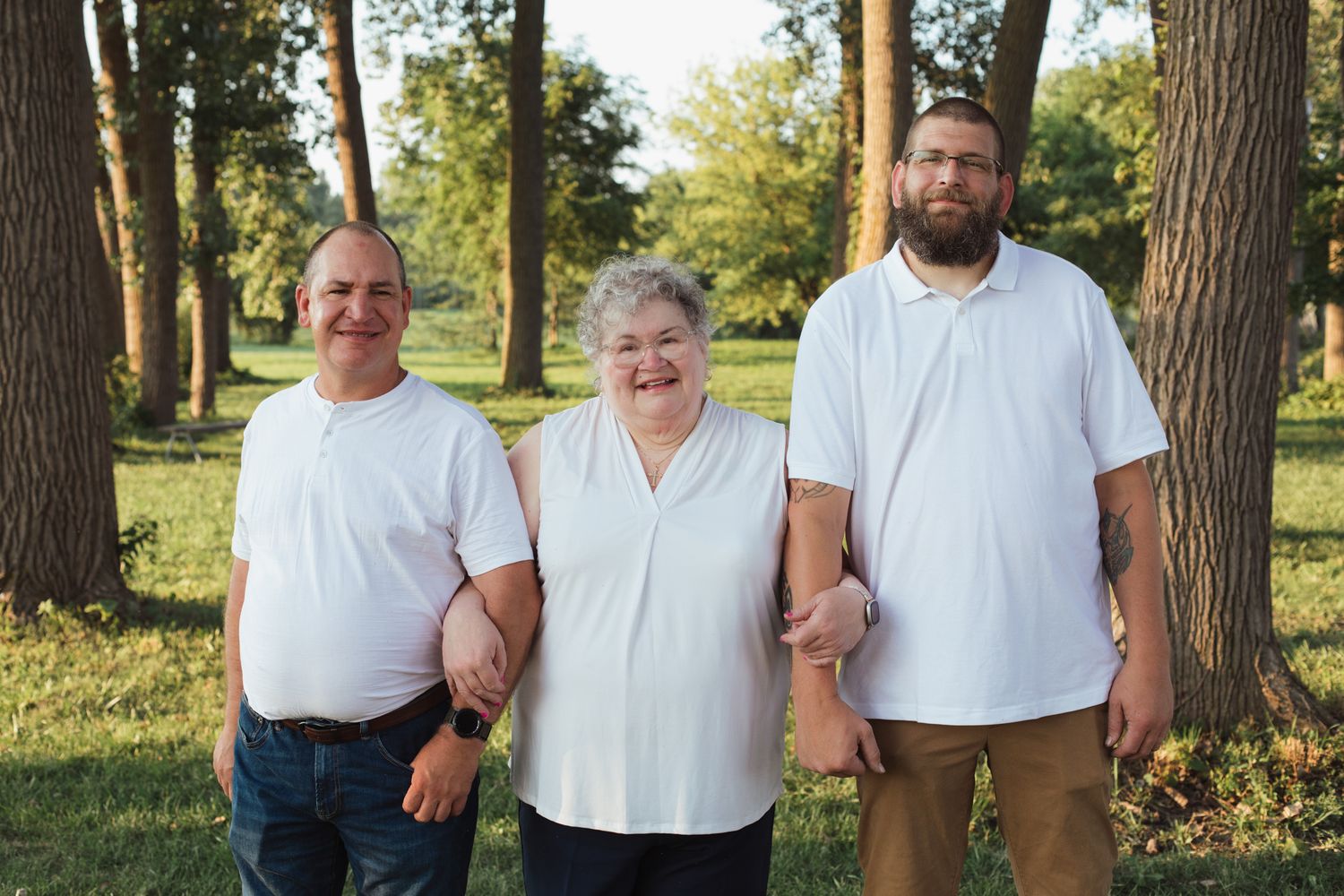 Three adults in white shirts smiling together outdoors in a lush green park on a sunny day.
