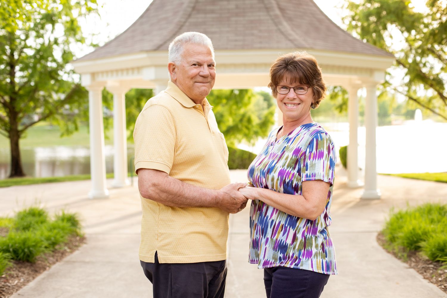 Smiling senior couple holding hands outdoors near a white gazebo in a lush green park setting.