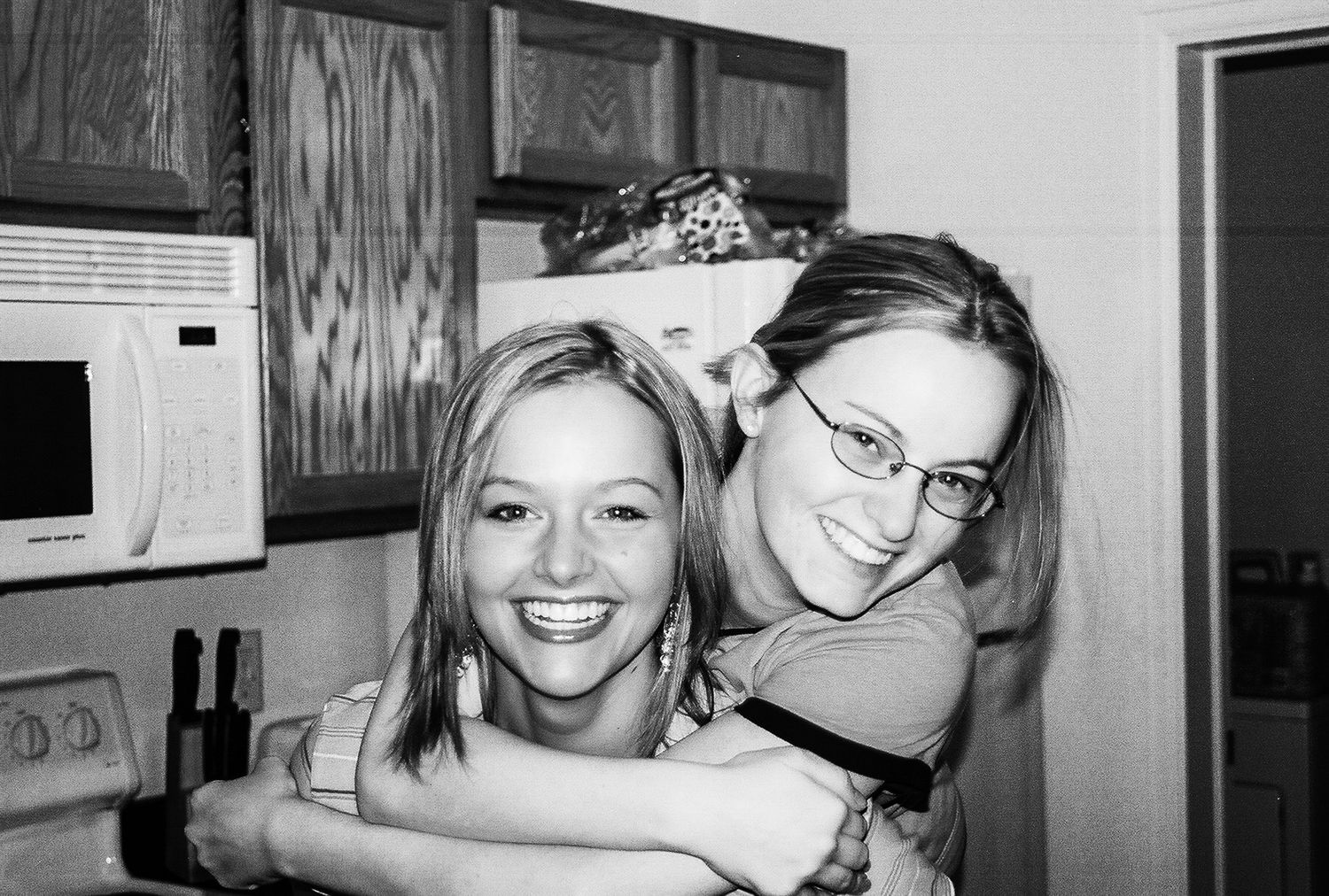 Two smiling young women hugging in a kitchen, black and white photo.