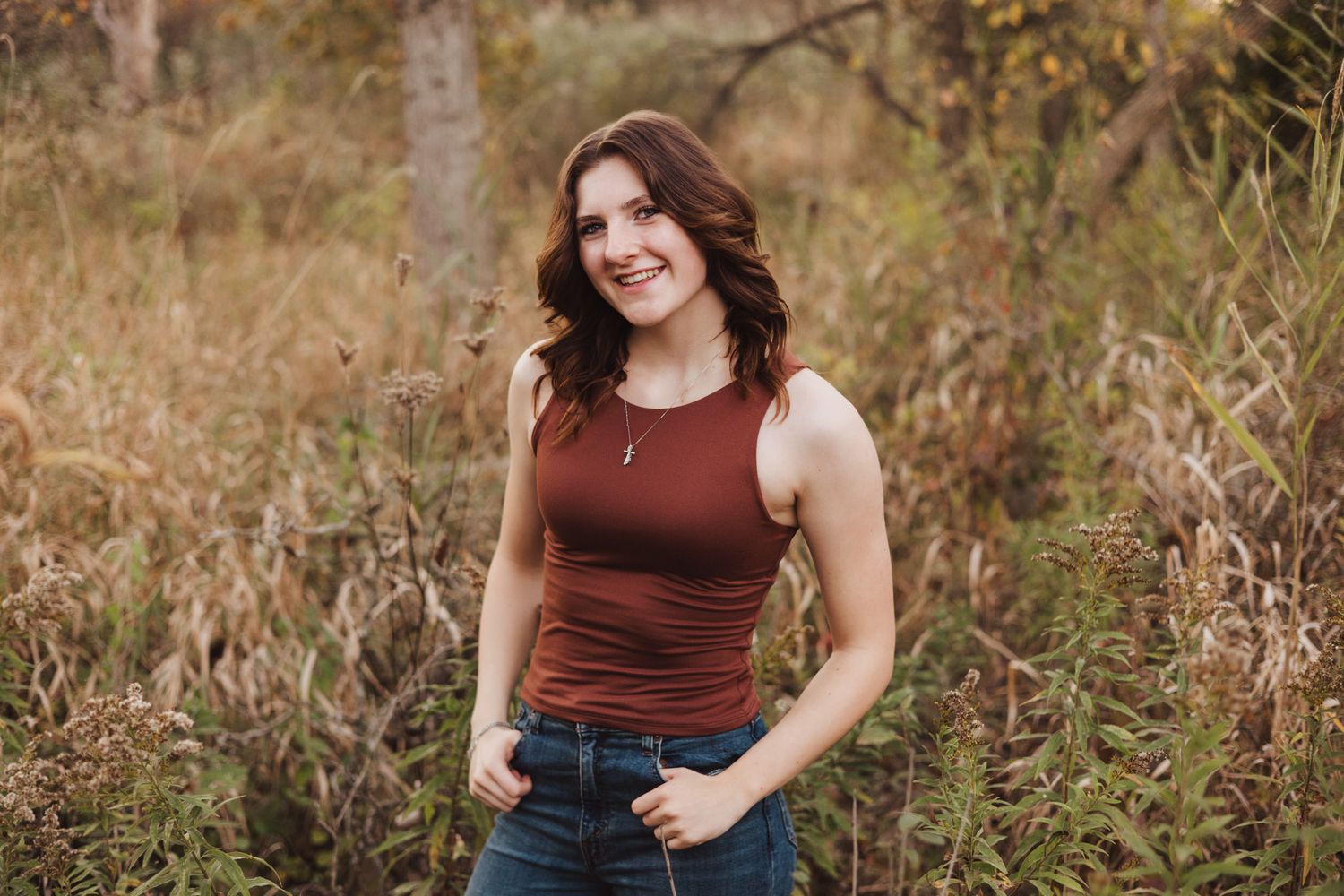 Young woman in red tank top and jeans smiling outdoors in autumn field with golden grasses.