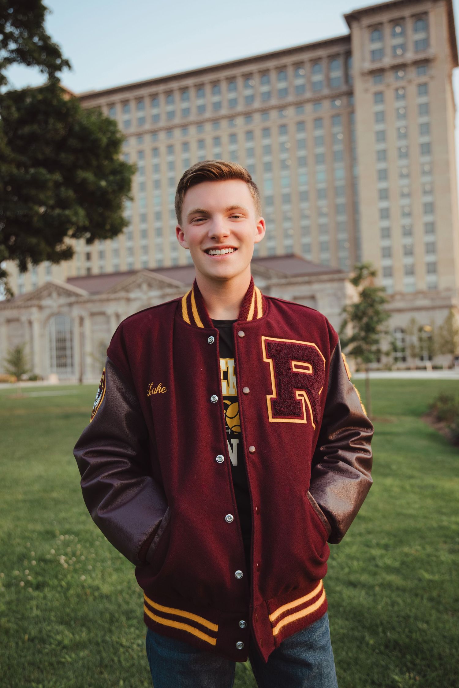 Smiling teen in maroon varsity jacket standing on lawn in front of historic building.