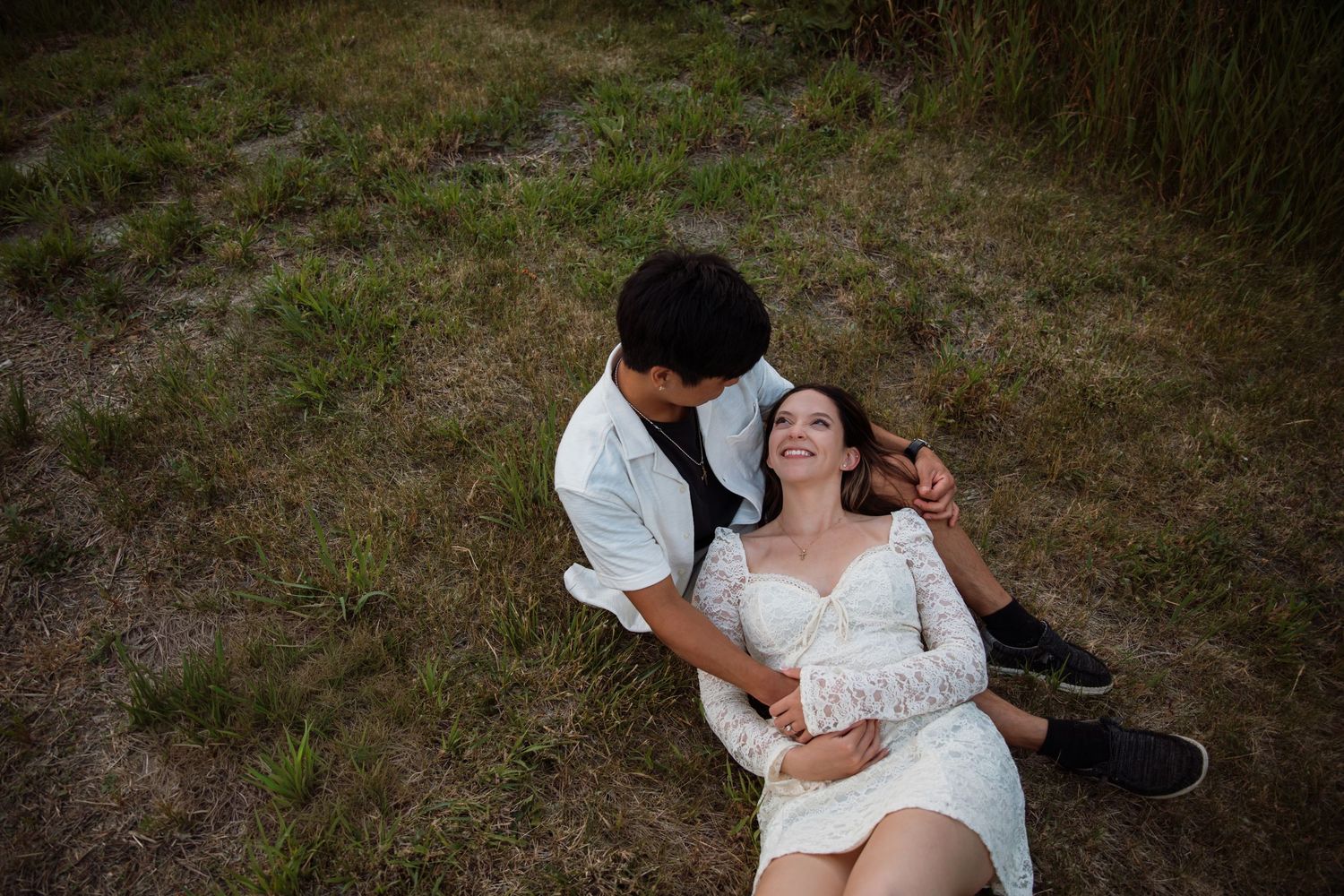 A joyful couple relaxes in a grassy field, with the woman laughing as her partner leans over her.