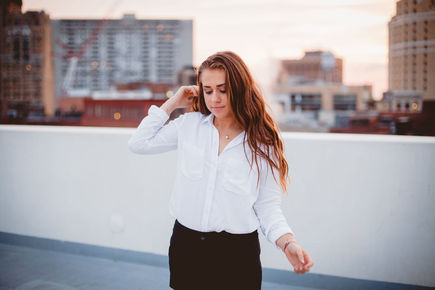 Young woman in white button-down shirt poses on rooftop with warm sunset city backdrop.
