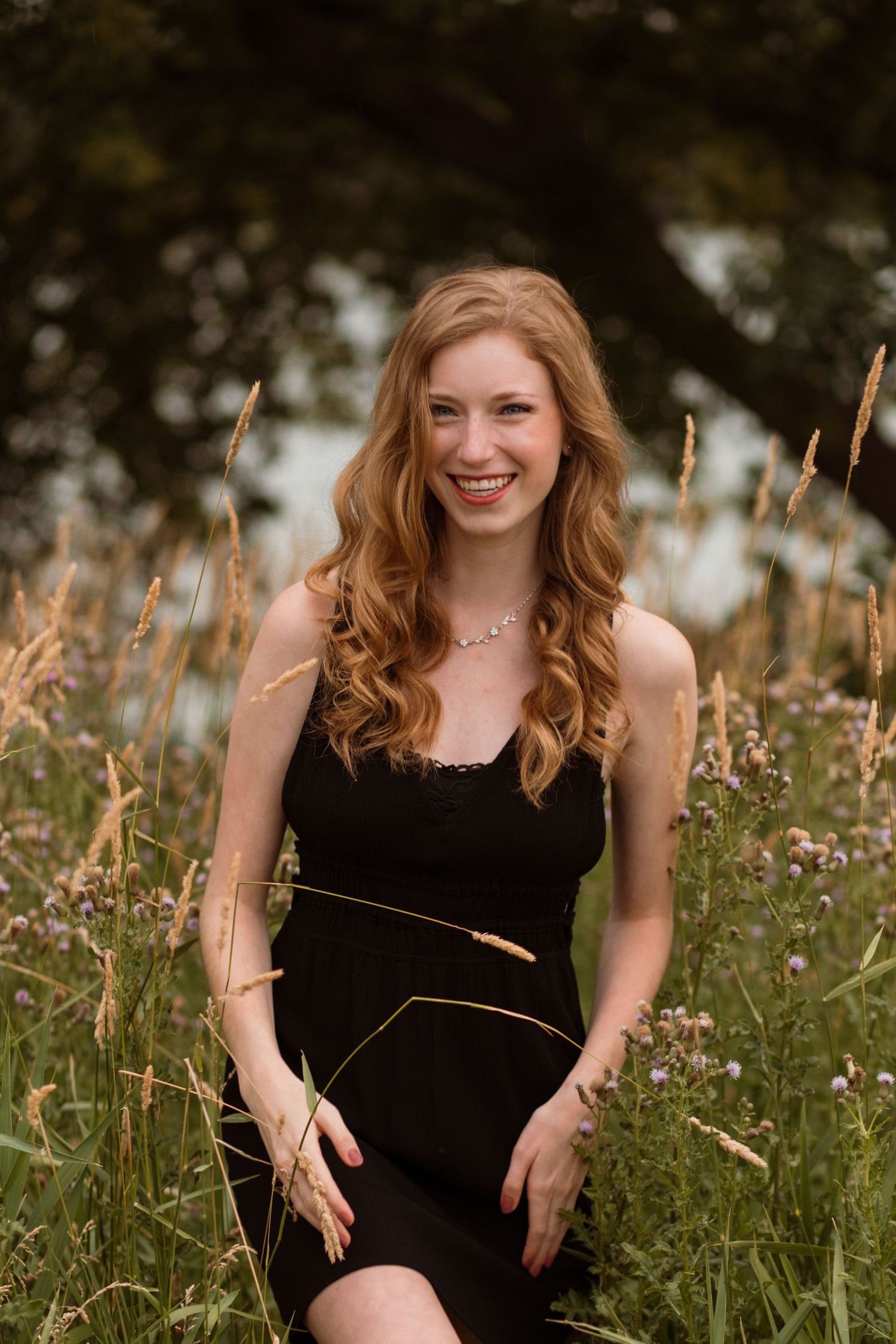 Smiling young woman in black dress sitting in wildflower meadow with tall grass and trees.