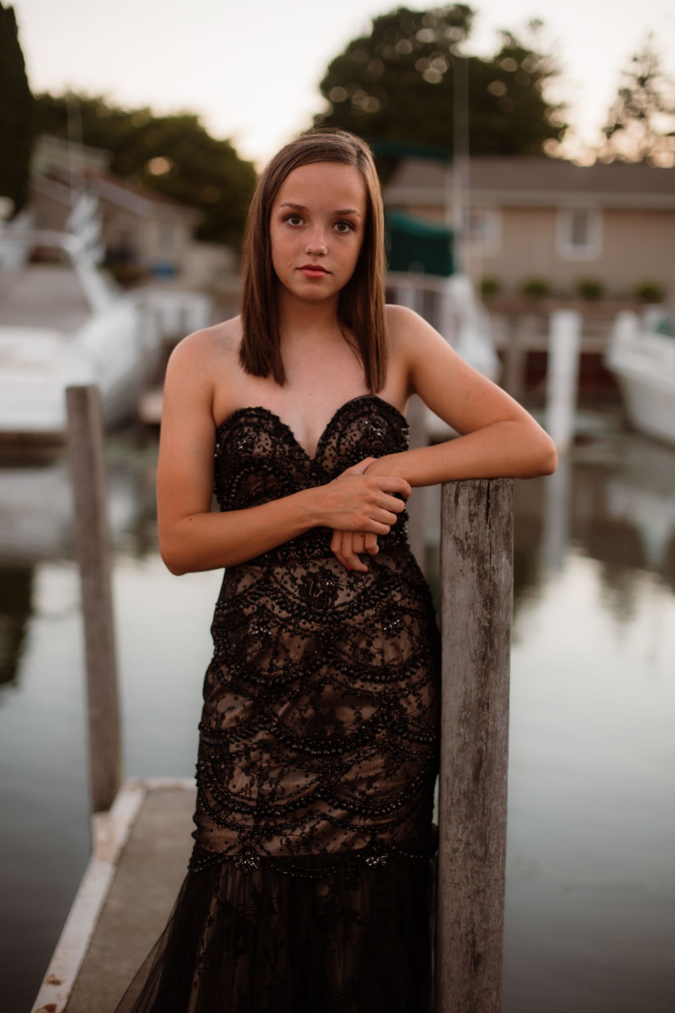 Teen girl in black lace mermaid gown leaning on wooden dock post at waterfront marina.