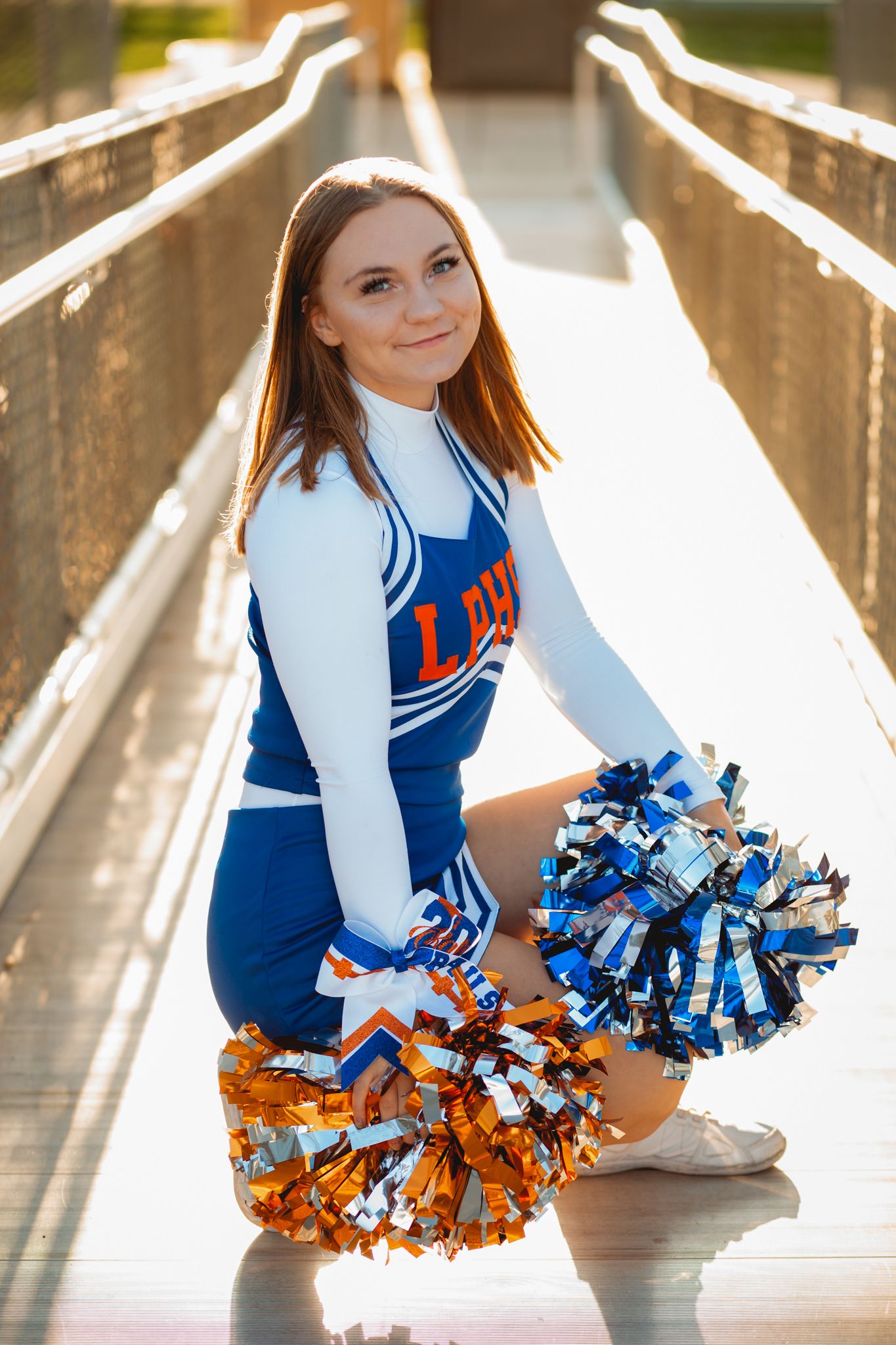 High school cheerleader in blue and white uniform kneeling with orange and silver pom-poms on bridge.