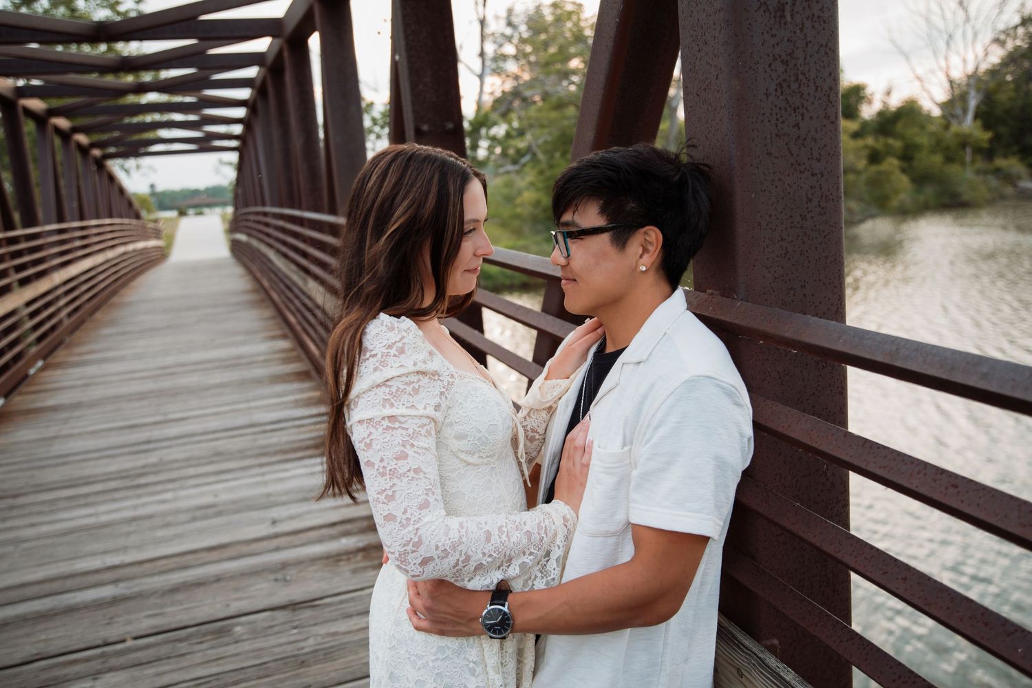 Couple embracing on a wooden bridge, woman in lace dress and man in white shirt, sharing a romantic moment outdoors.