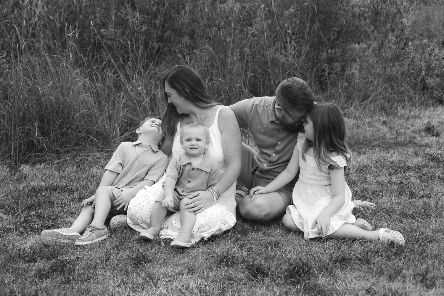 Black and white family portrait of parents and three young children sitting together on grass outdoors.