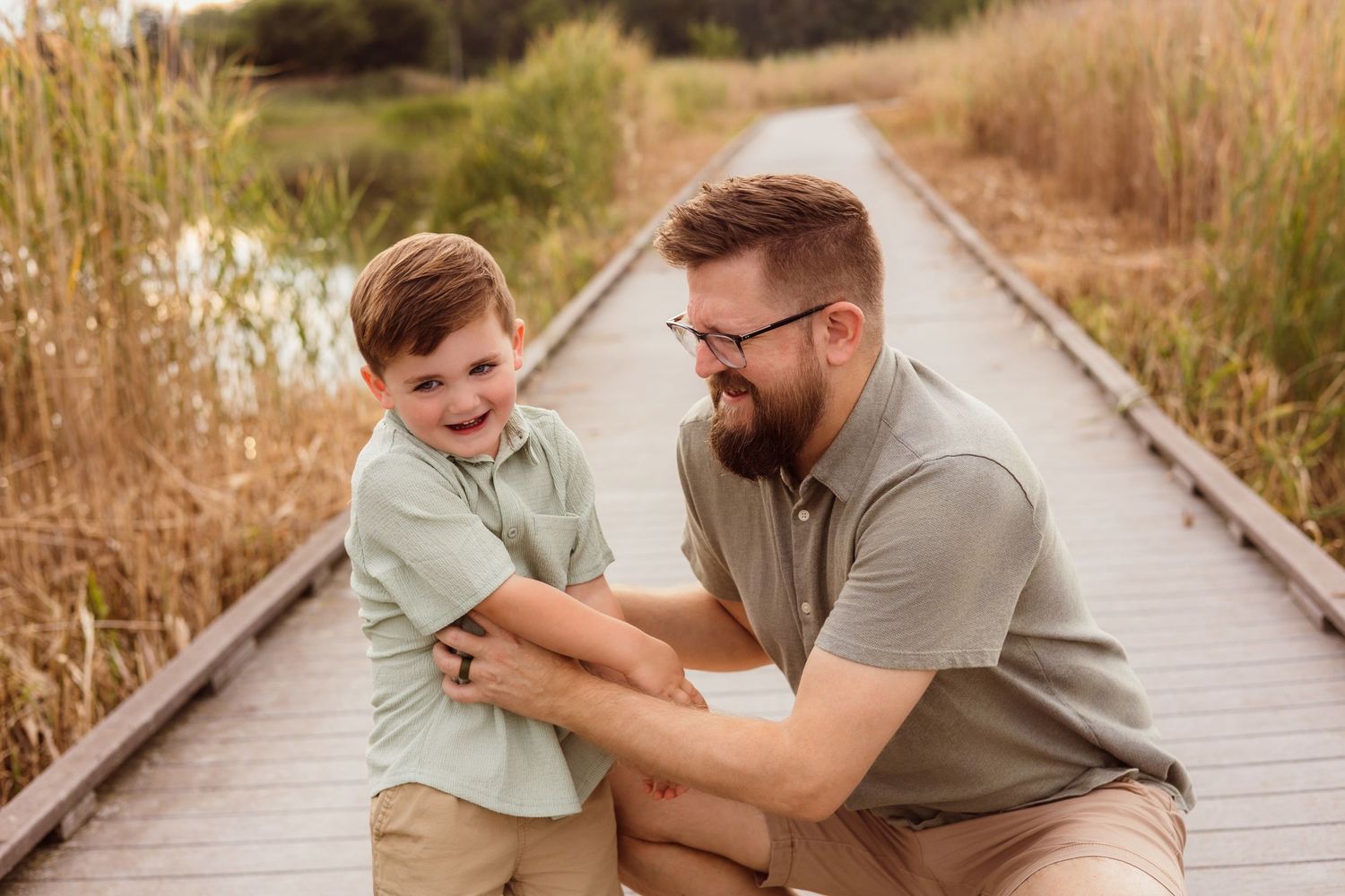 A father and young son laugh together on a wooden boardwalk path surrounded by tall golden grass.