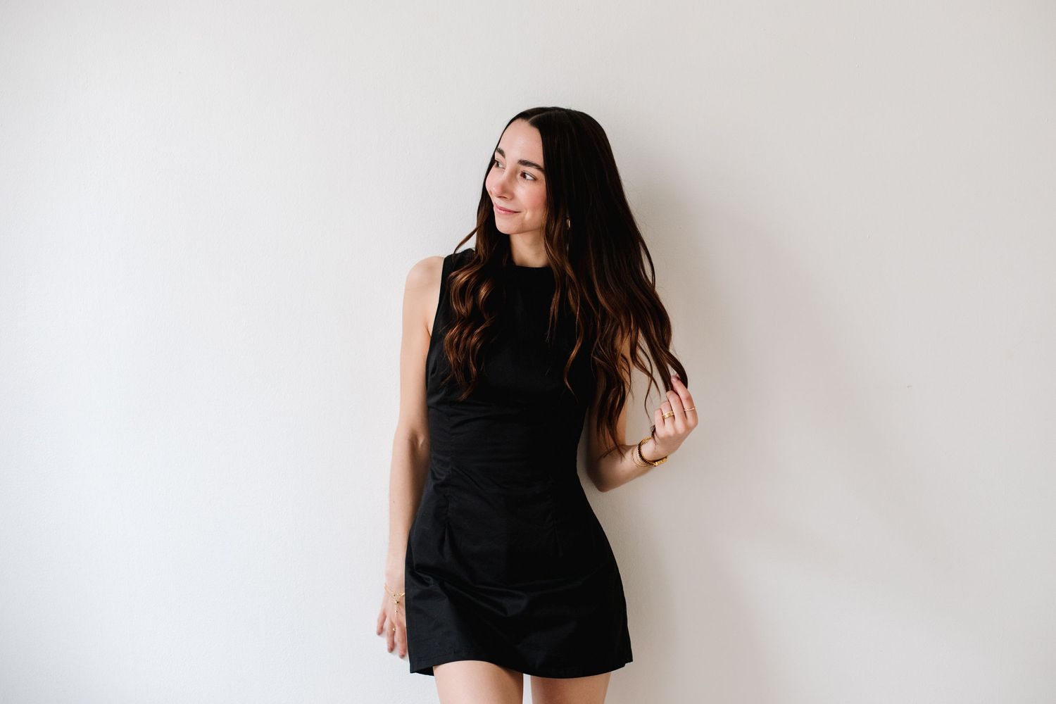 Young woman with long dark hair in black sleeveless mini dress posing against white wall.