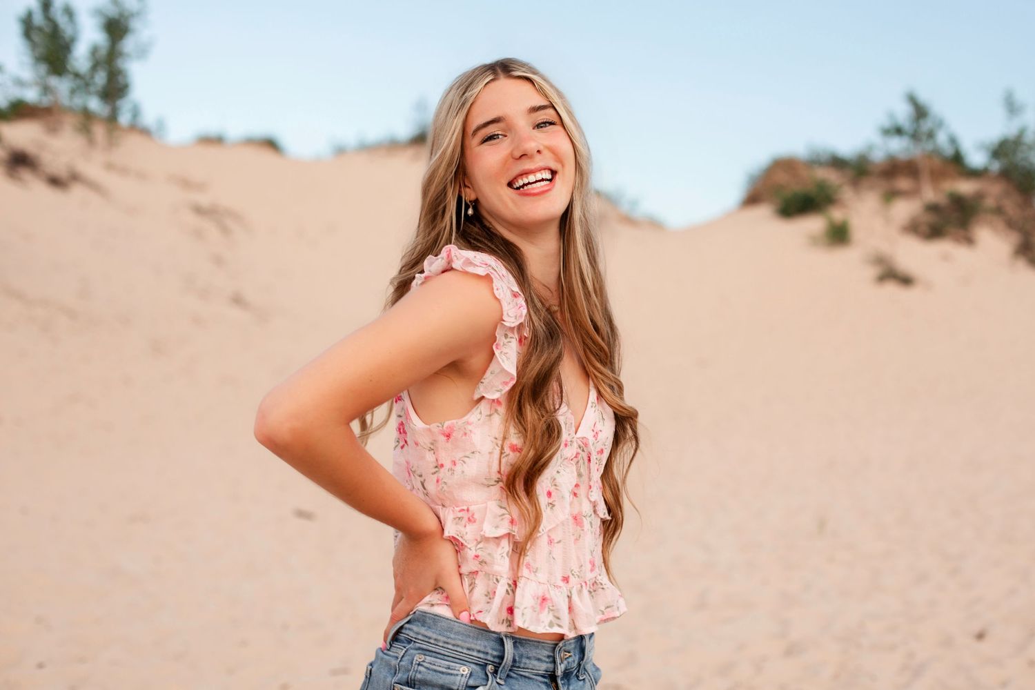 Smiling young woman in floral top and jeans posing on sandy dunes under a clear blue sky.