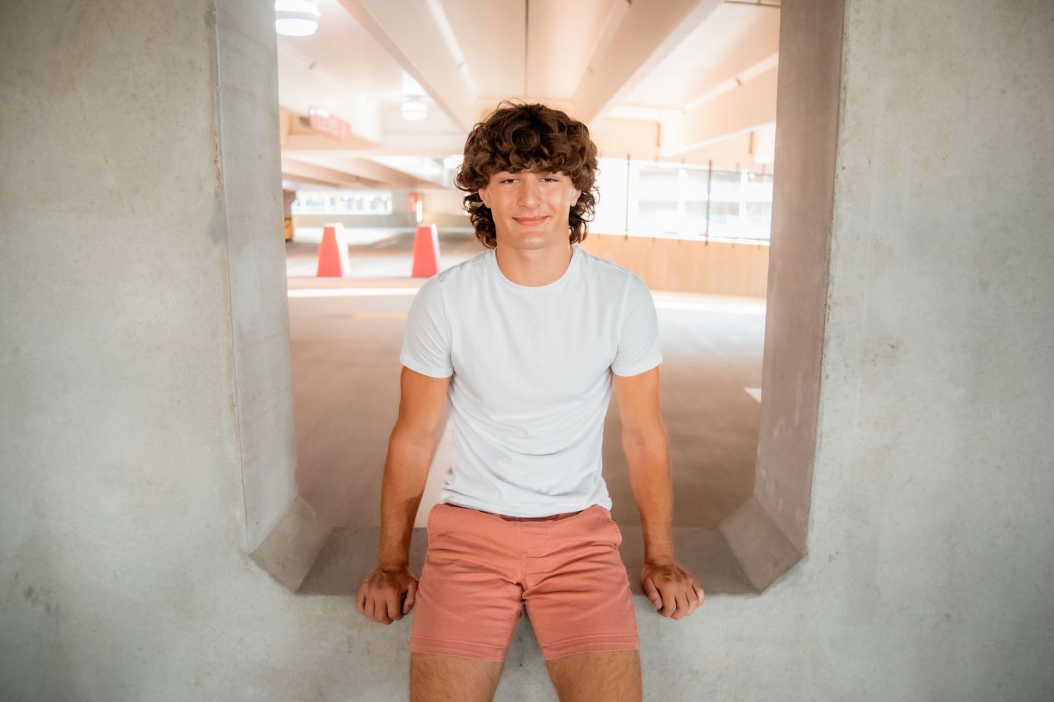 Young man with curly hair wearing white t-shirt and pink shorts sitting in a parking garage.
