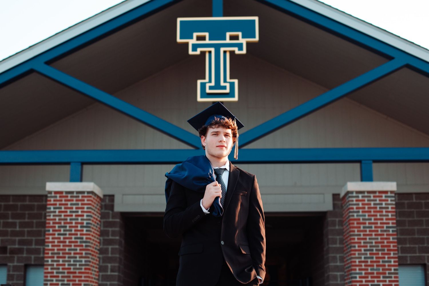 A high school graduate in cap and gown poses confidently in front of a brick school entrance with a blue T logo.