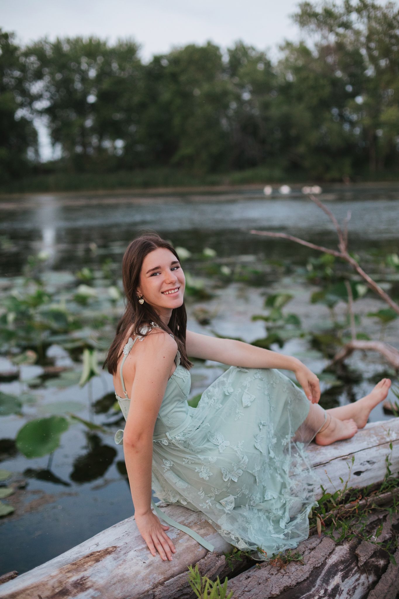 Smiling young woman in mint green dress sitting on a log by a lily pad pond.