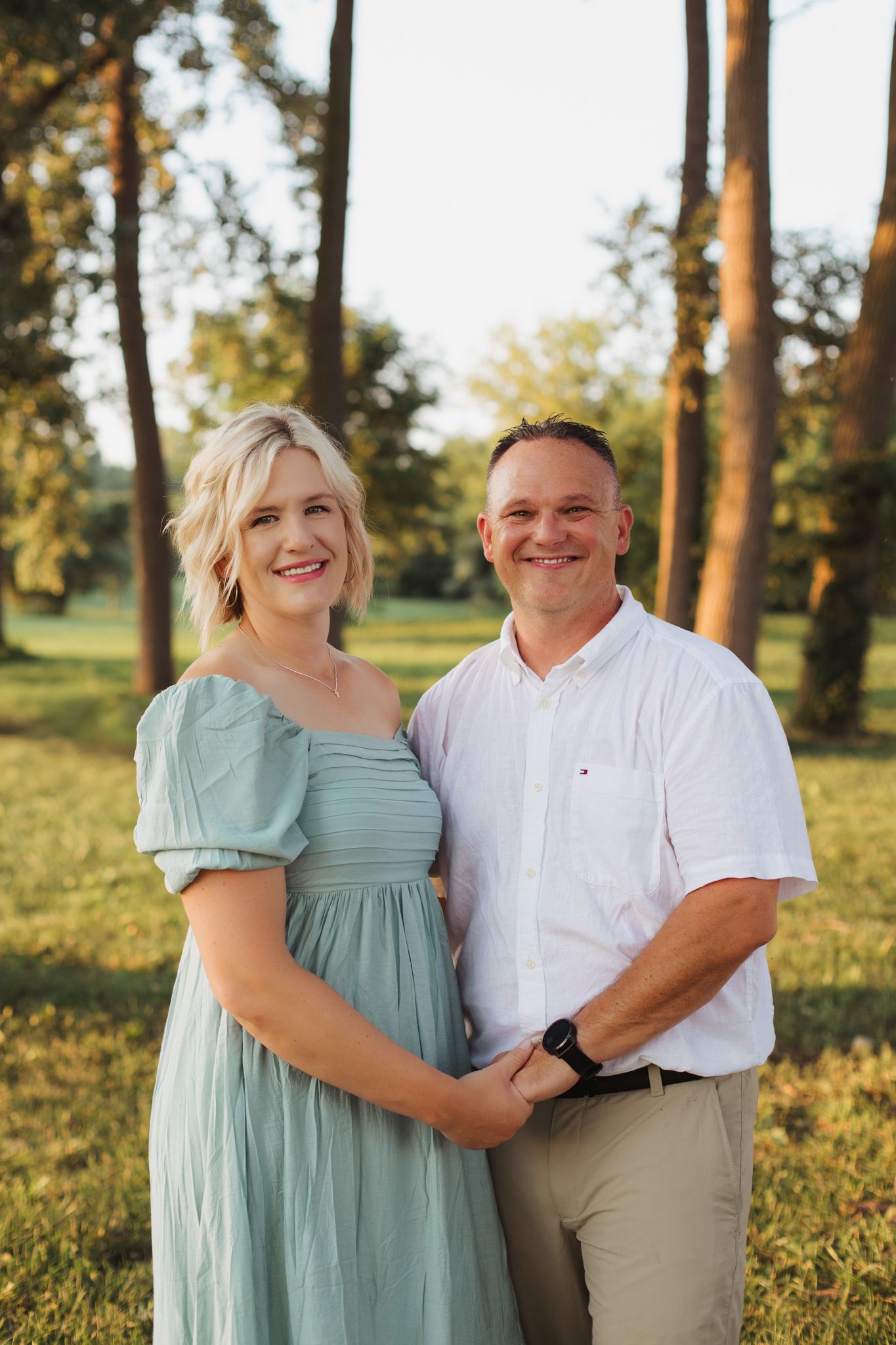 A smiling couple poses outdoors in golden sunlight, woman in sage green dress, man in white shirt, trees in background.