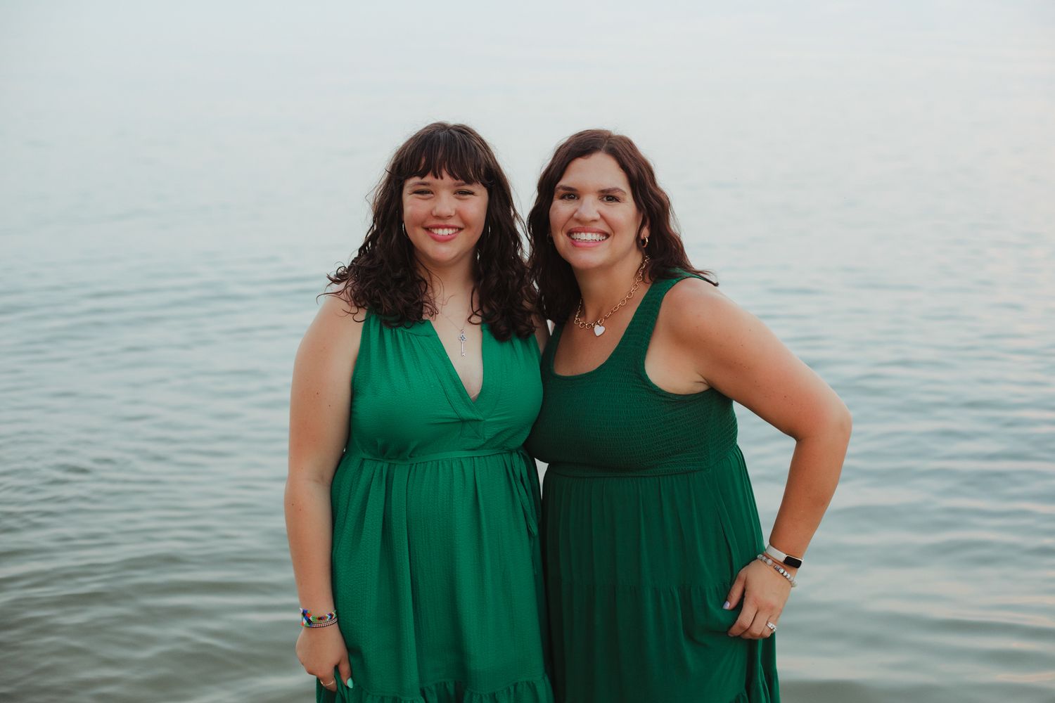 Two women in matching green dresses smiling together near a calm body of water at dusk.