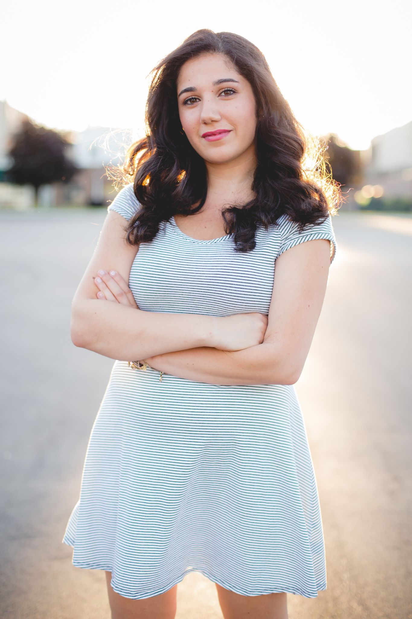 Young woman in striped dress standing outdoors with arms crossed, smiling confidently in warm golden sunlight.
