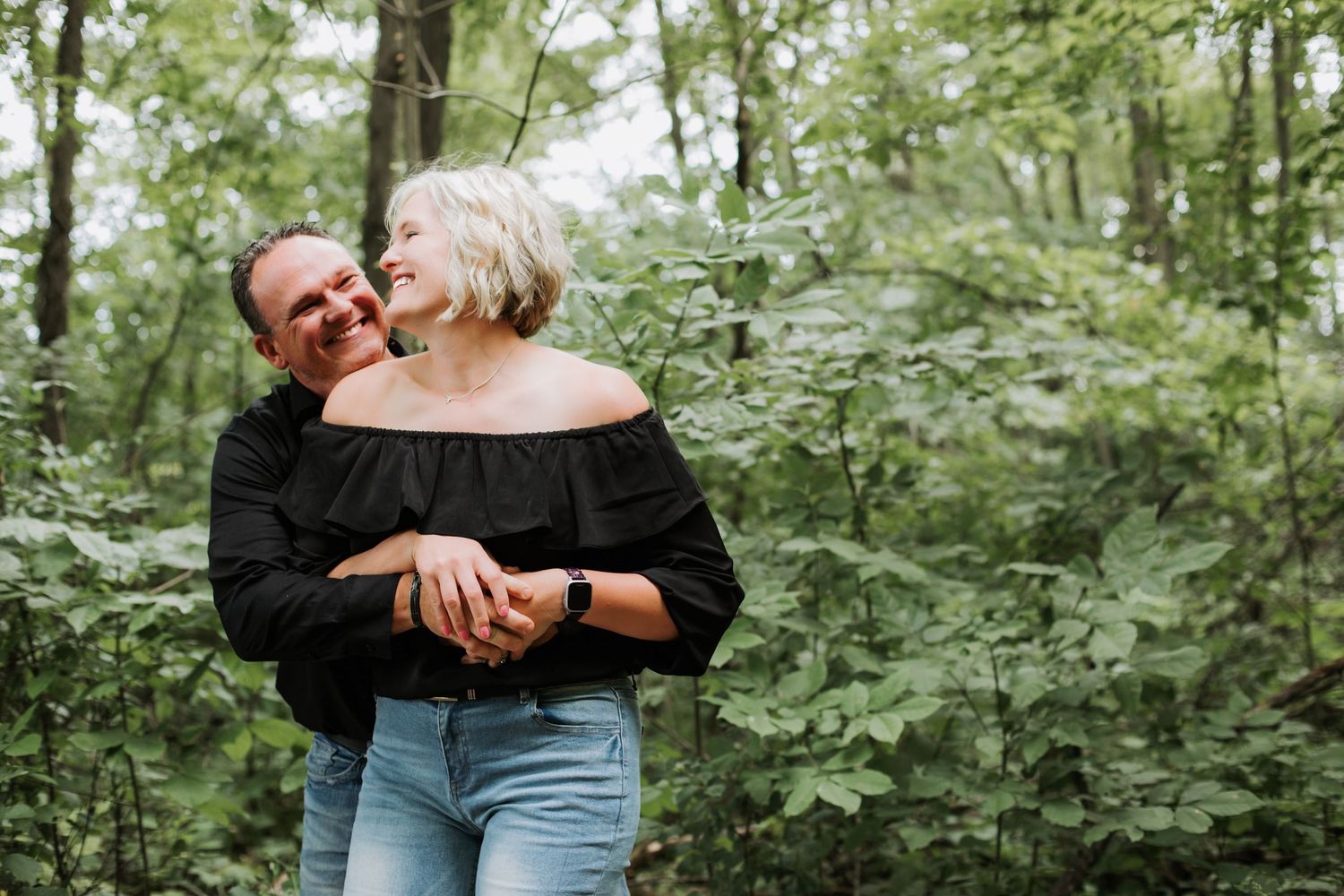 A happy couple in black tops share a warm embrace while laughing together in a lush green forest setting.