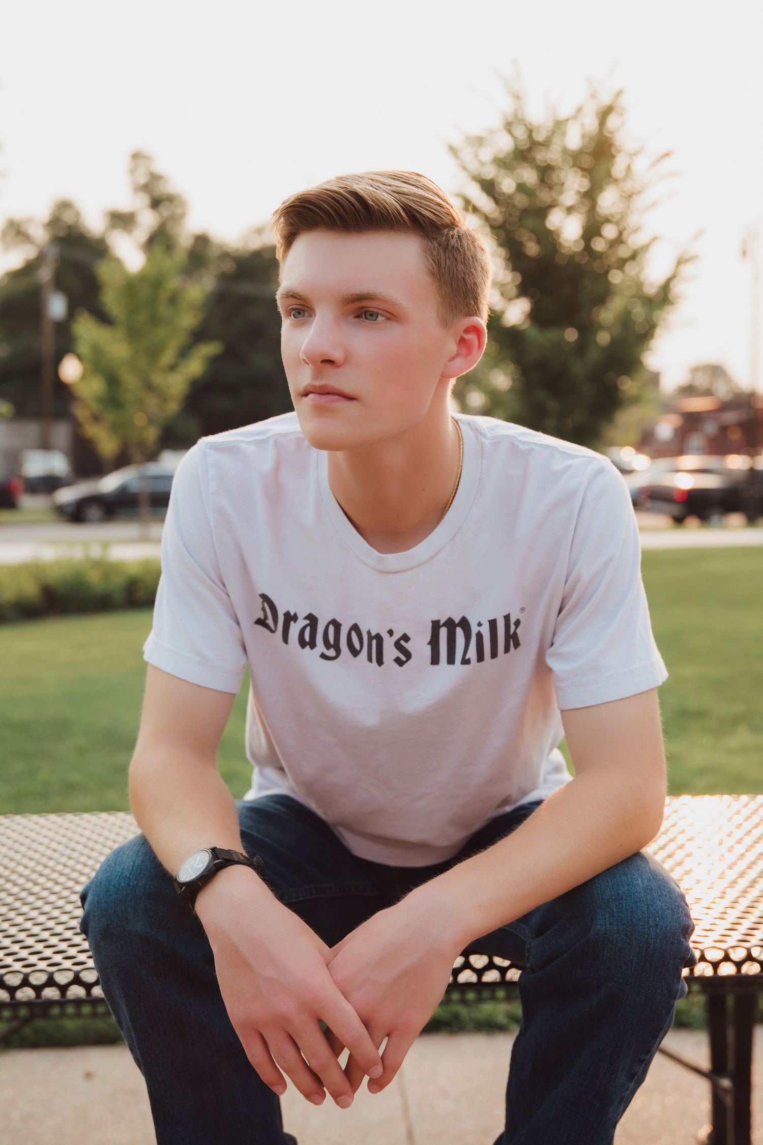 Young man in Dragon's Milk white t-shirt sitting on bench in park at golden hour.