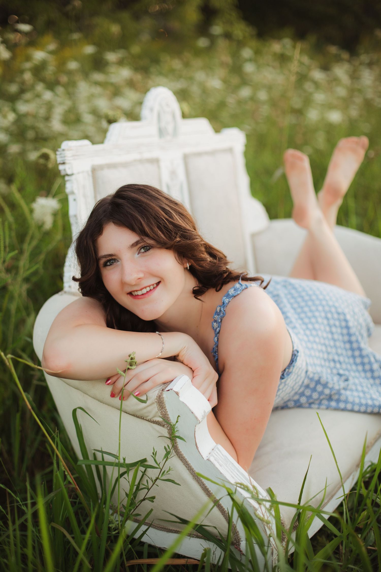 Happy young woman in blue gingham dress posing on white antique chair in wildflower field.