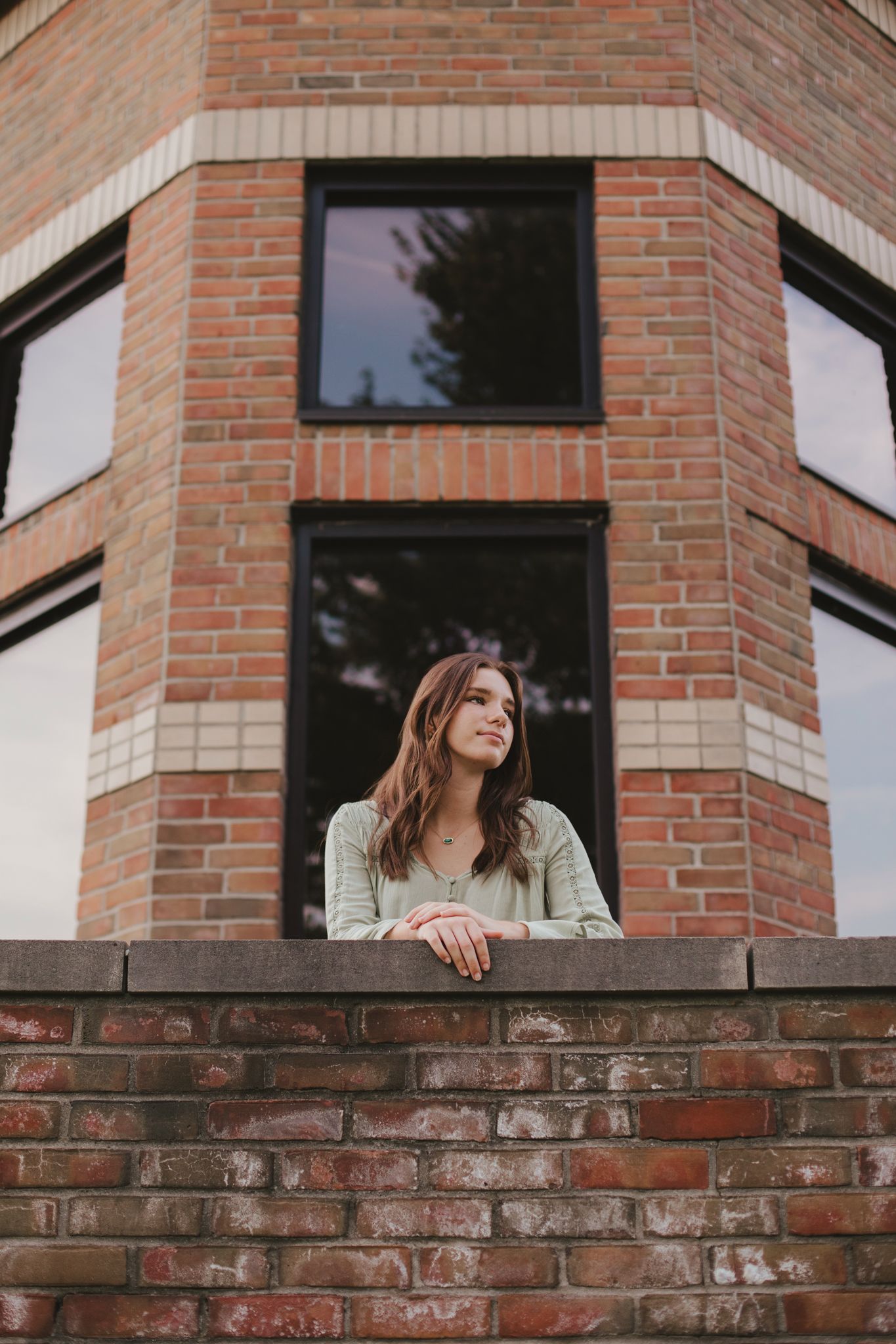 Young woman in sage green top leaning on brick wall, gazing thoughtfully beside a modern red brick building.