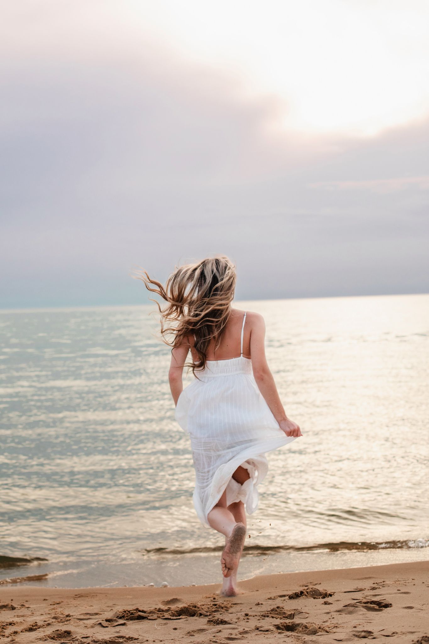 Woman in white dress running barefoot on sandy beach toward calm ocean water at sunset.