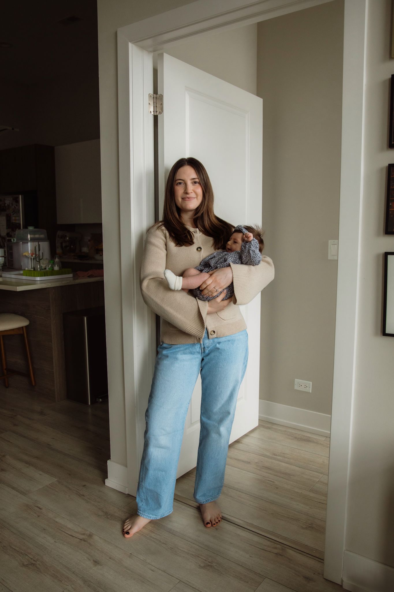 Smiling mother in beige cardigan and jeans holding newborn baby in home doorway.