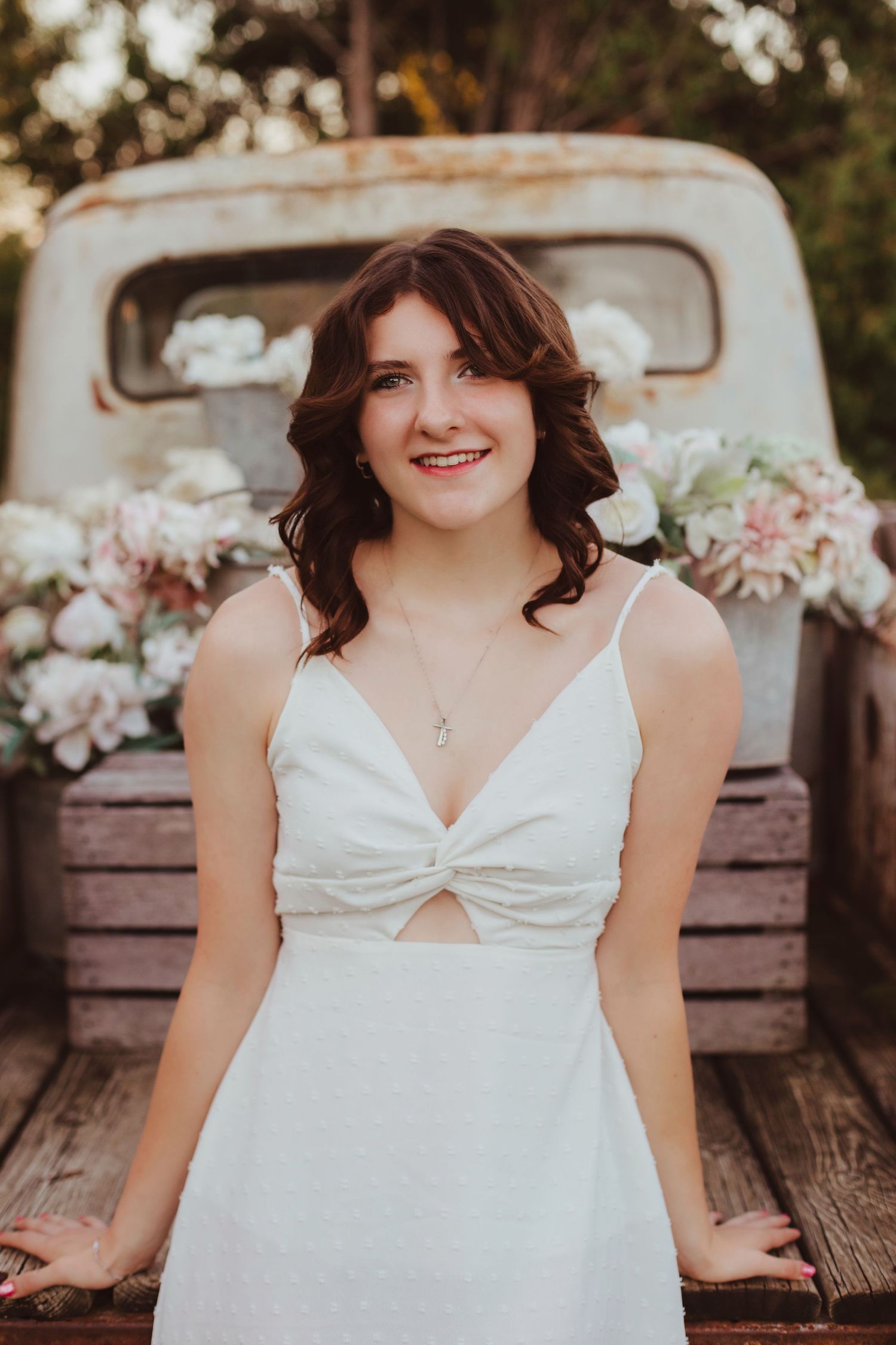 Smiling young woman in white dress standing at a vintage truck bed decorated with pink and white flowers.