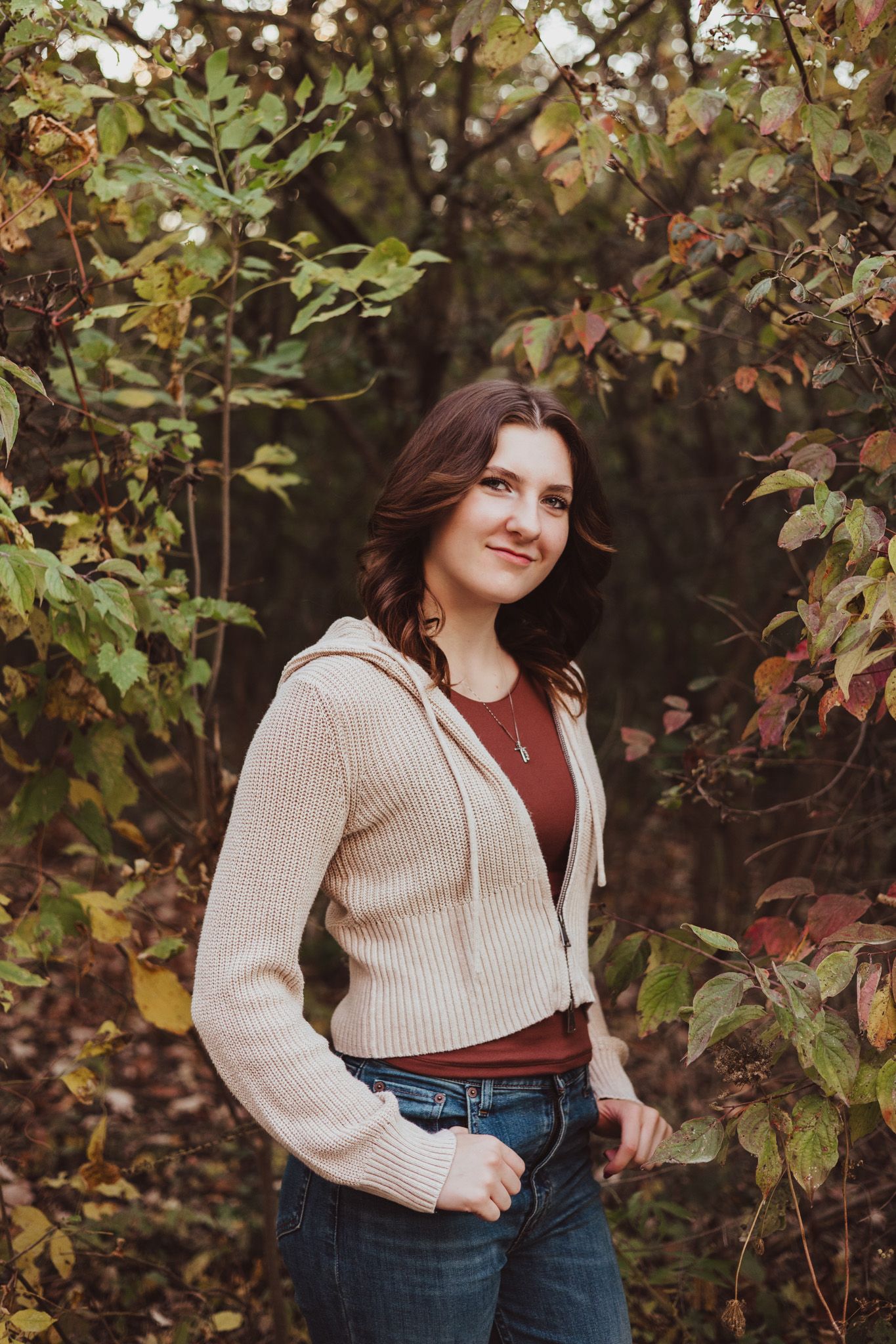 Young woman in cream hoodie and jeans poses among colorful autumn foliage in a forest setting.