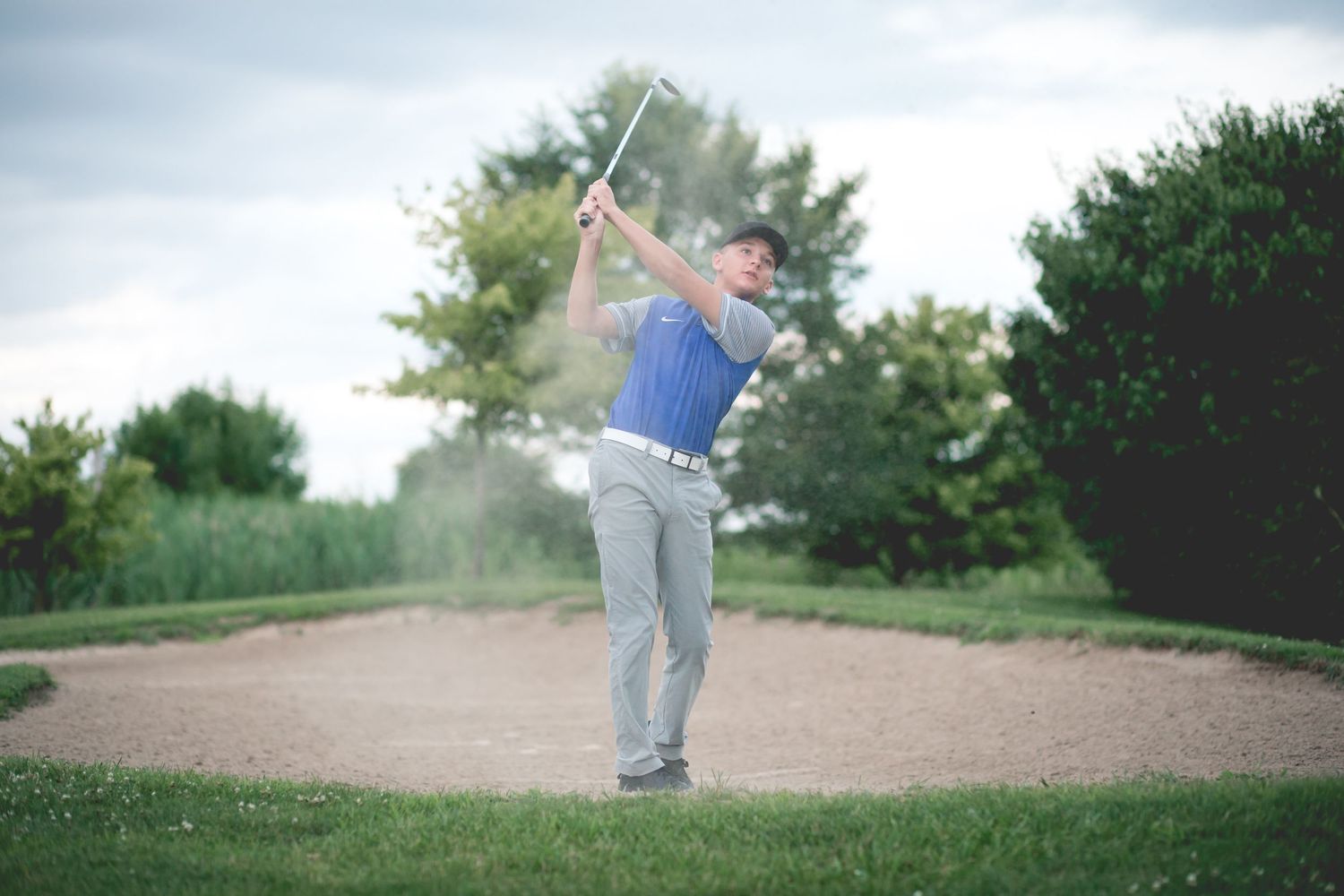 Golfer hitting a sand bunker shot on a golf course, sending sand flying with trees in the background.