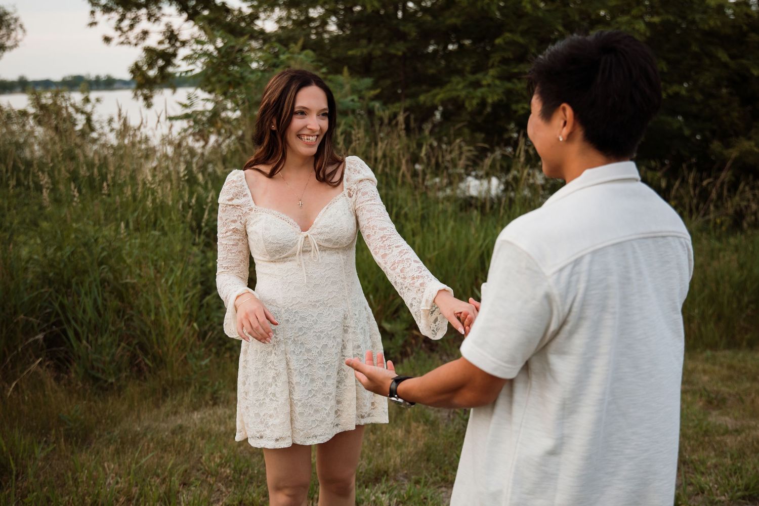 Smiling woman in white lace dress holds hands with partner near a scenic lakeside meadow.