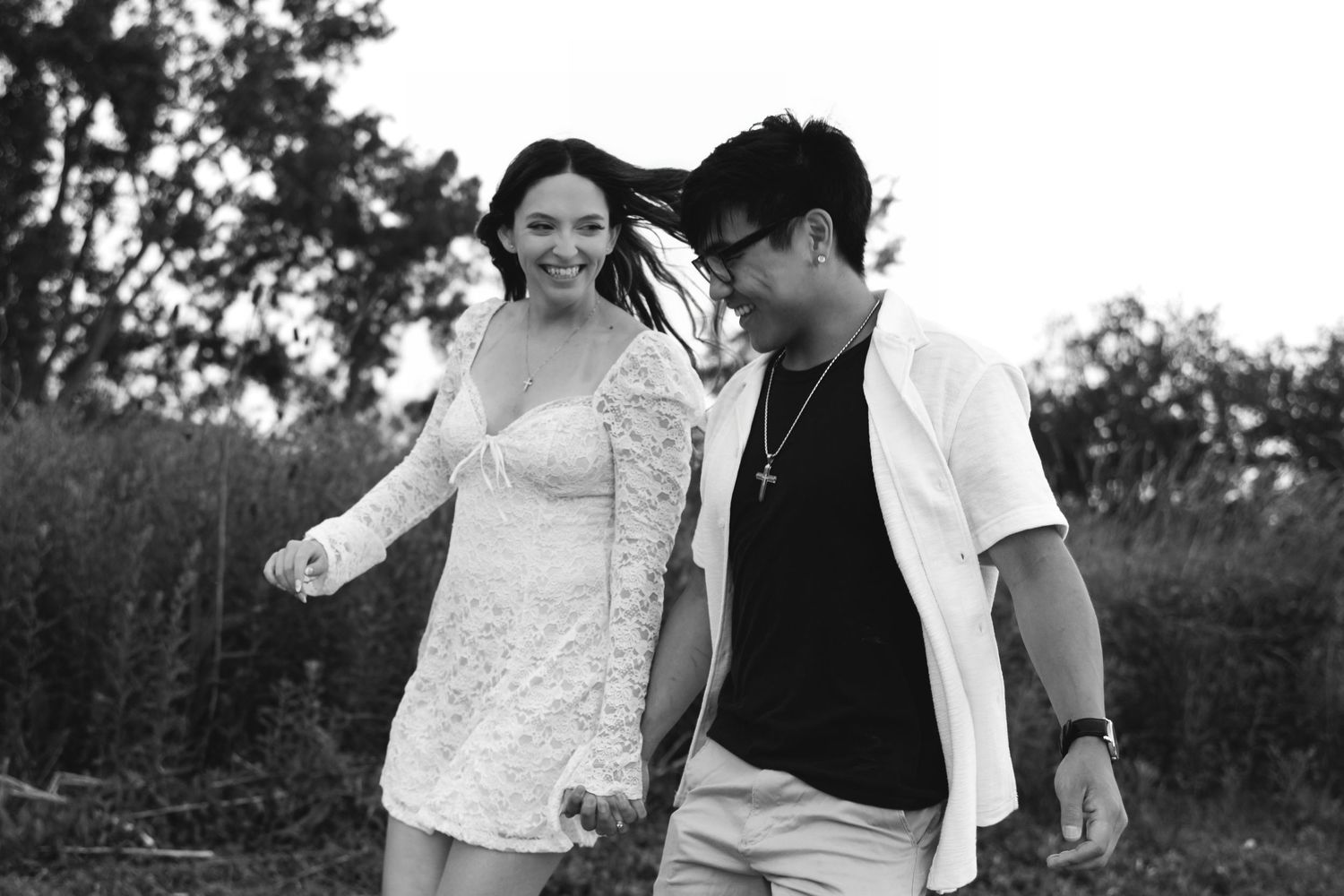 Black and white photo of a smiling couple walking outdoors near trees and tall grass on a windy day.