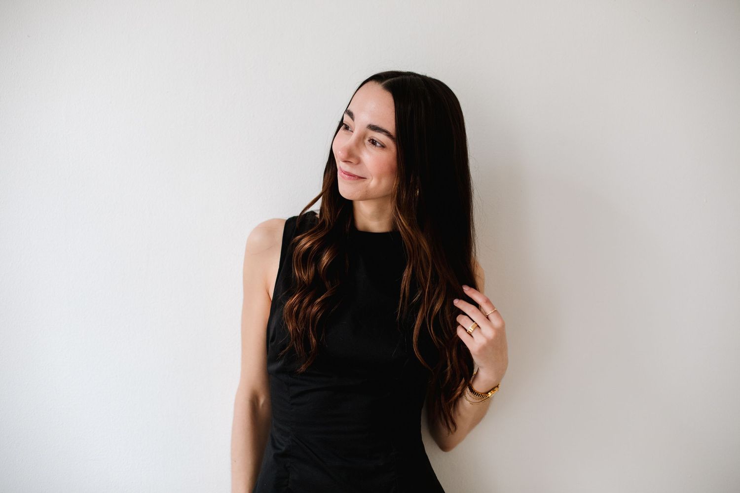 Brunette woman in black sleeveless dress gazing sideways against neutral white wall.