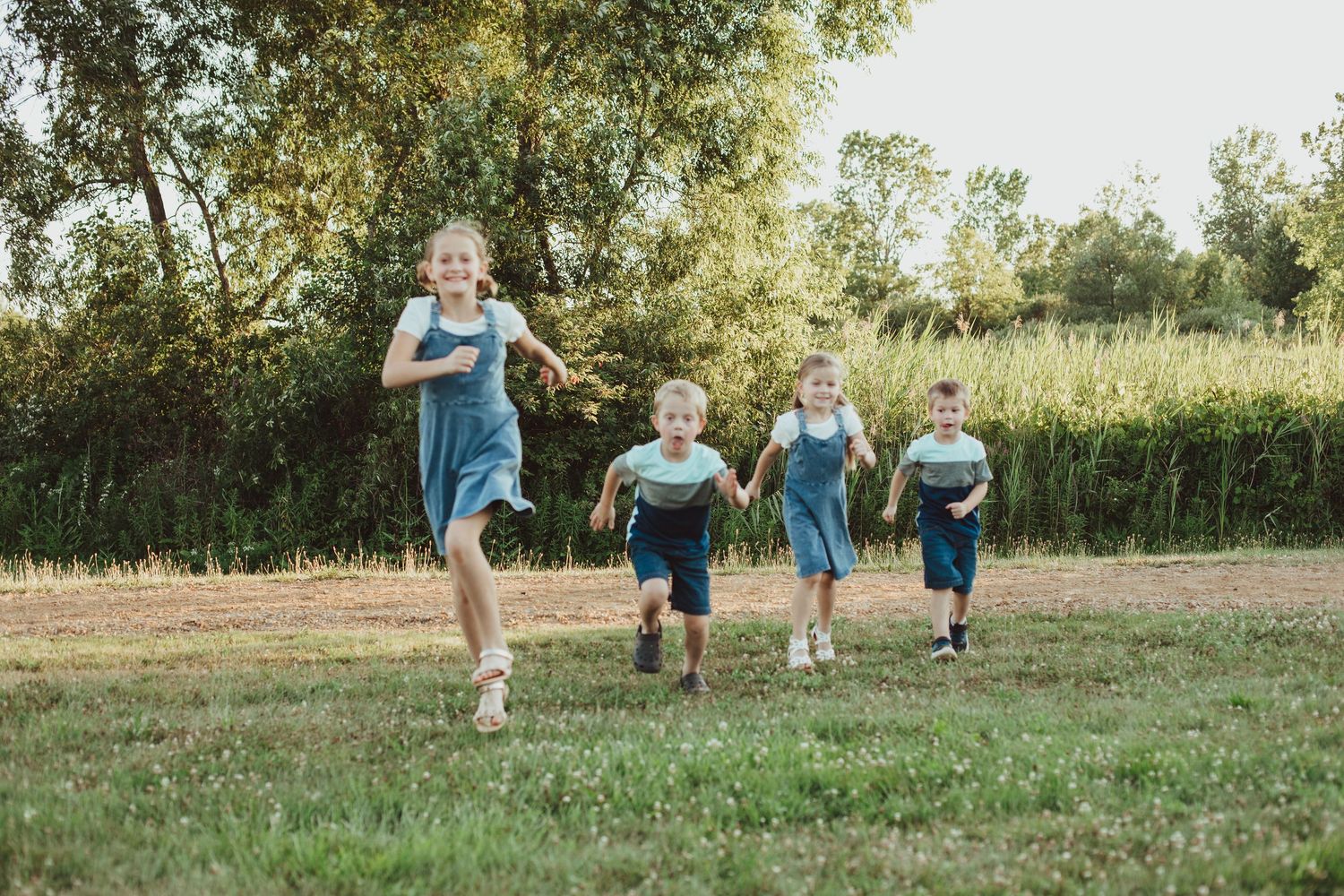 Four children in denim outfits running joyfully across a green field with trees in the background.