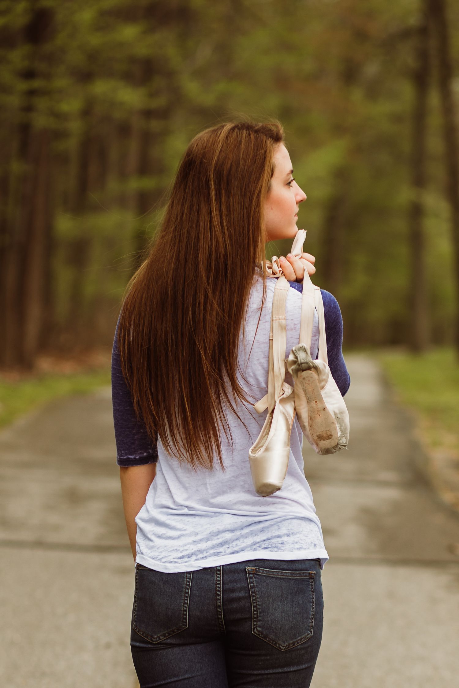 Young woman with long brown hair carrying ballet shoes while walking down forest road.