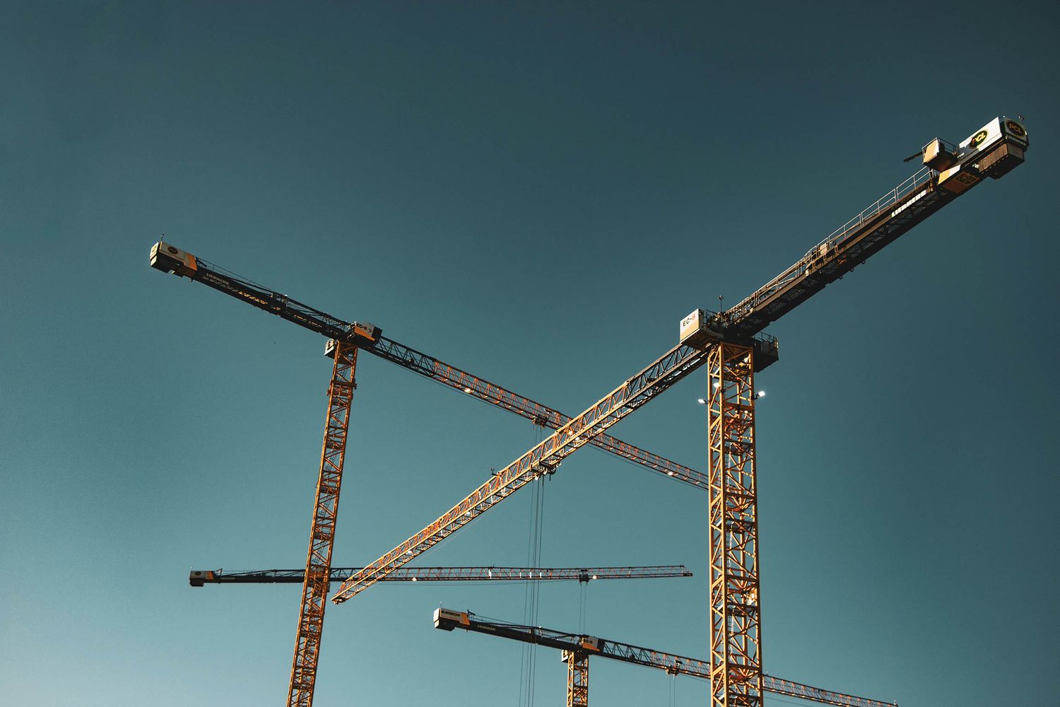 Four yellow construction cranes intersecting against a clear teal sky at a building site.