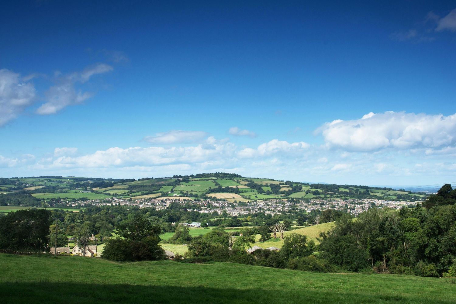 Panoramic view of a lush green valley town under blue skies with rolling hills in the British countryside.